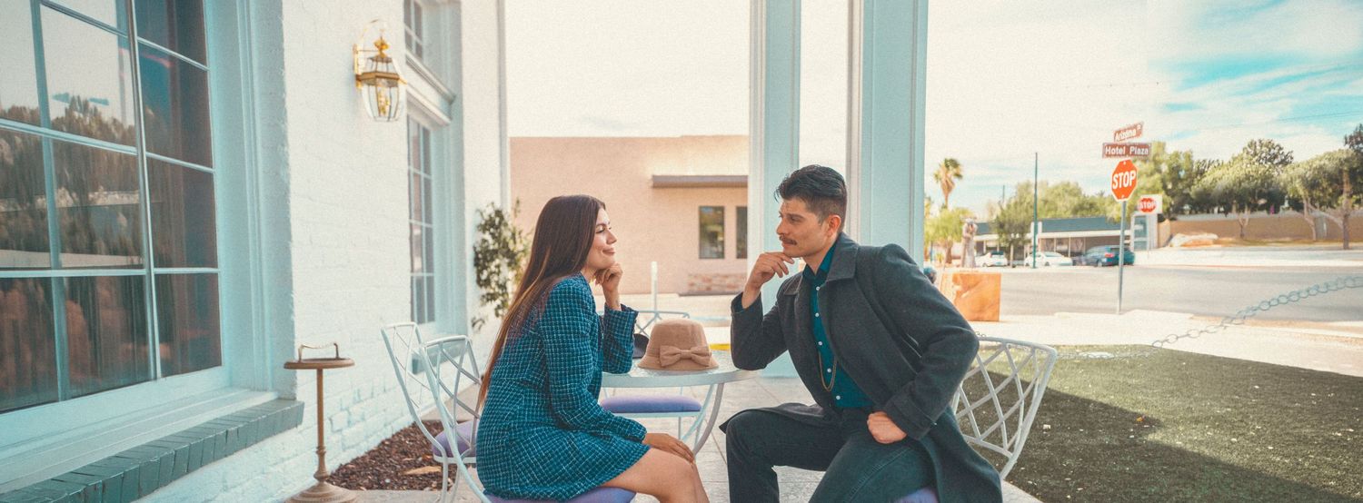 A couple sits at an outdoor cafe table, sharing a joyful moment while holding hands on a sunny day.