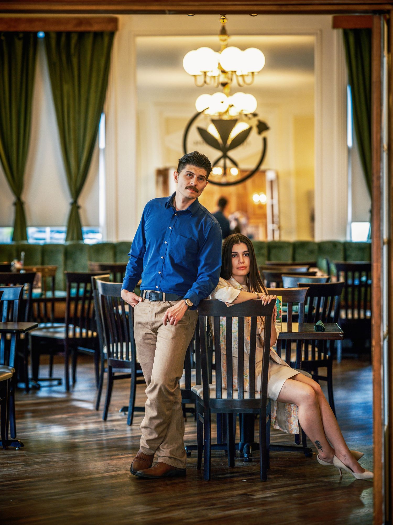 A man and woman pose elegantly in a upscale restaurant with wood floors, chandeliers, and green velvet seating.