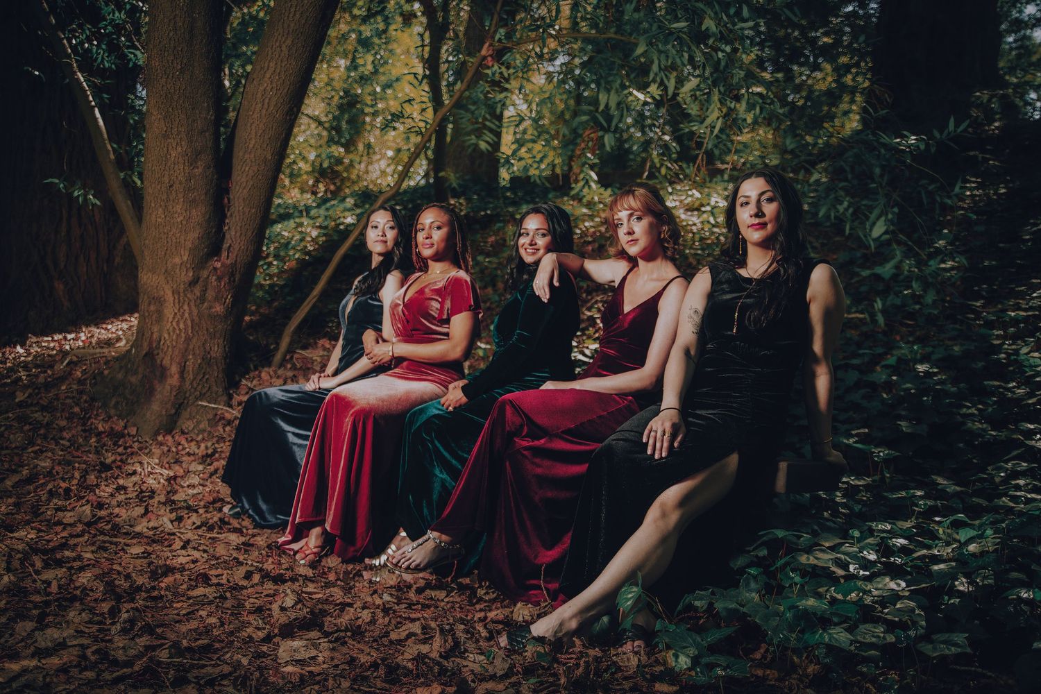 Five women in elegant dresses pose together in a moody forest setting surrounded by autumn leaves.