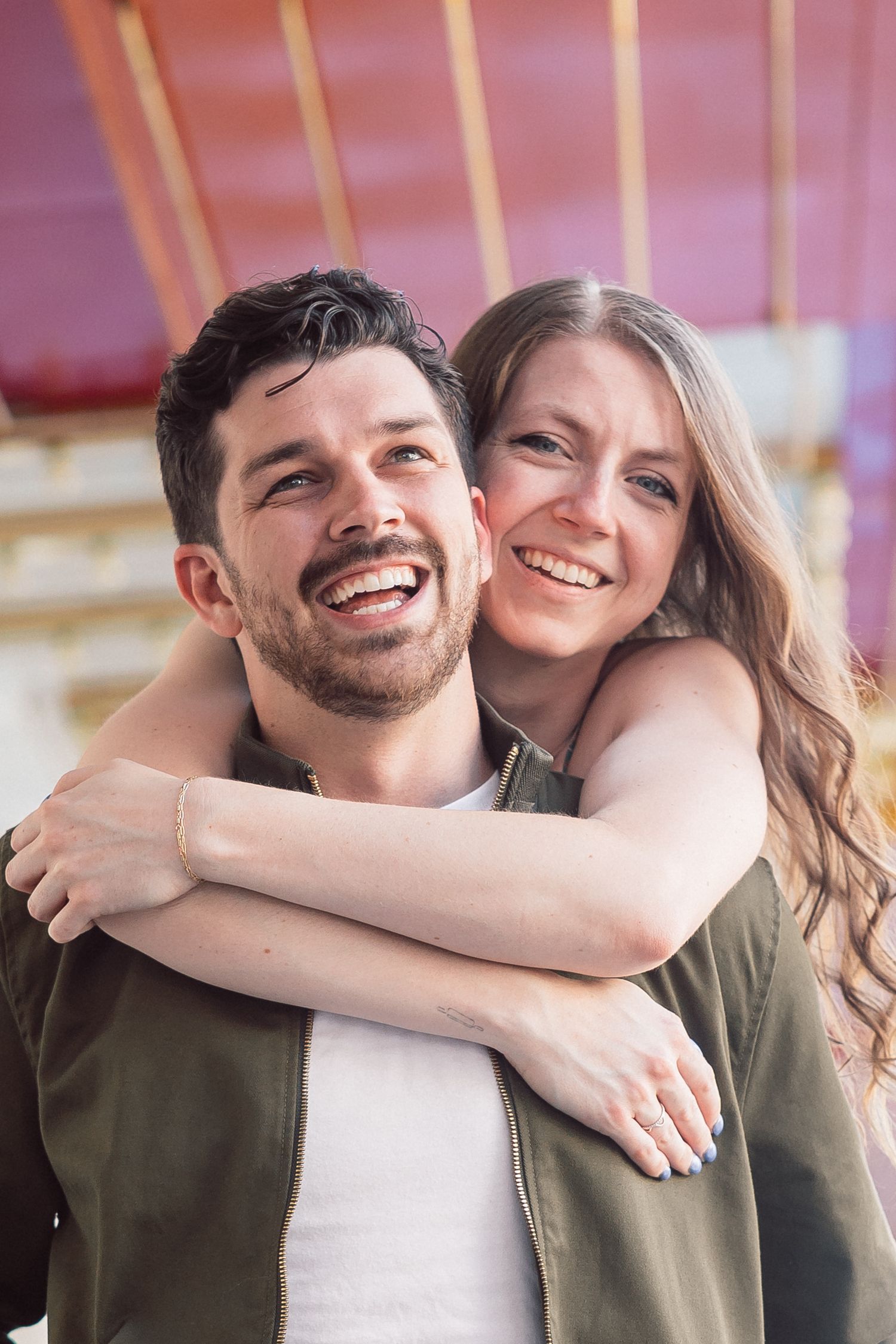 Happy couple smiling outdoors, woman hugging man from behind, both wearing green jackets, colorful background.