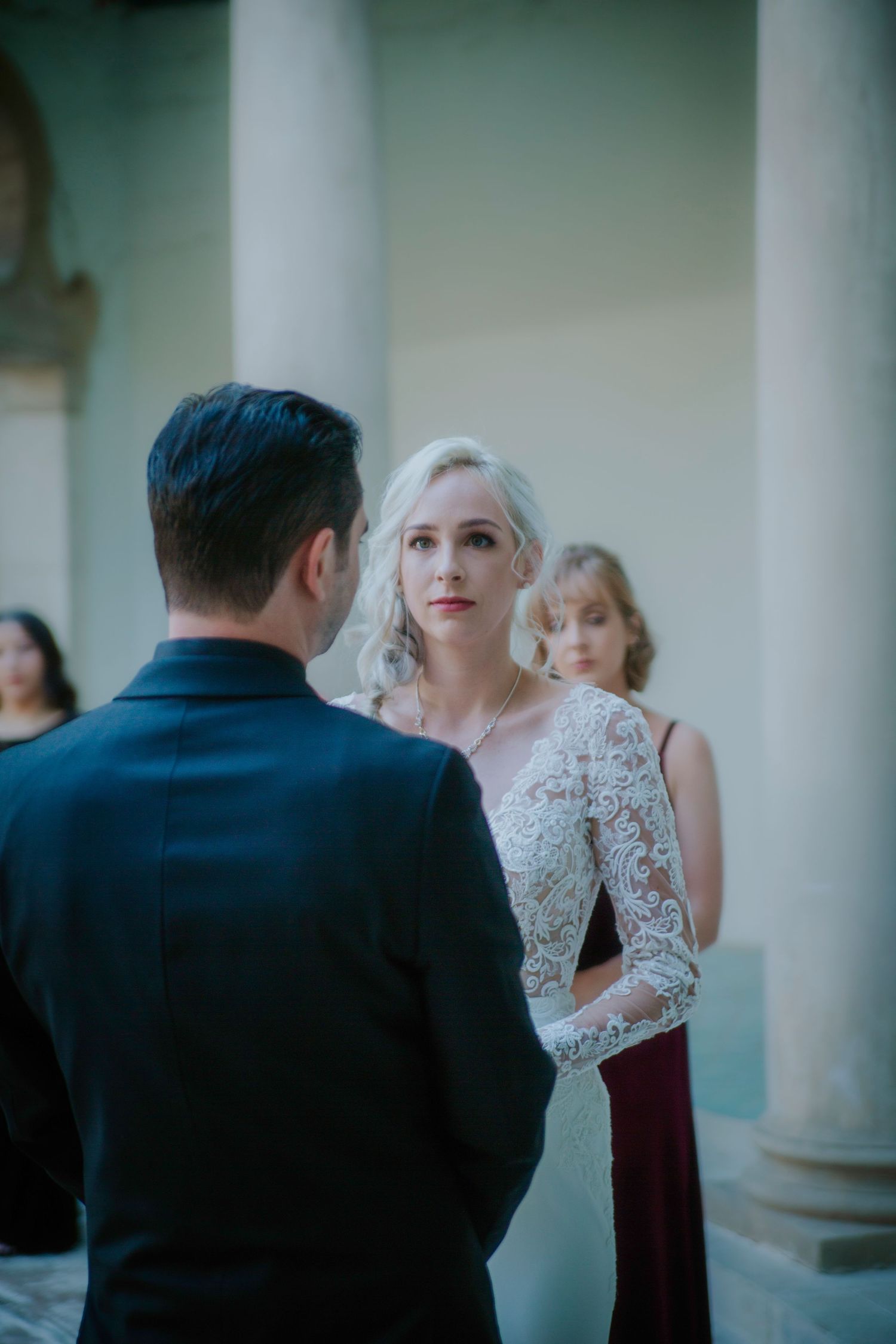 Bride in lace wedding dress gazes at groom during outdoor wedding ceremony with bridesmaid in burgundy dress behind.