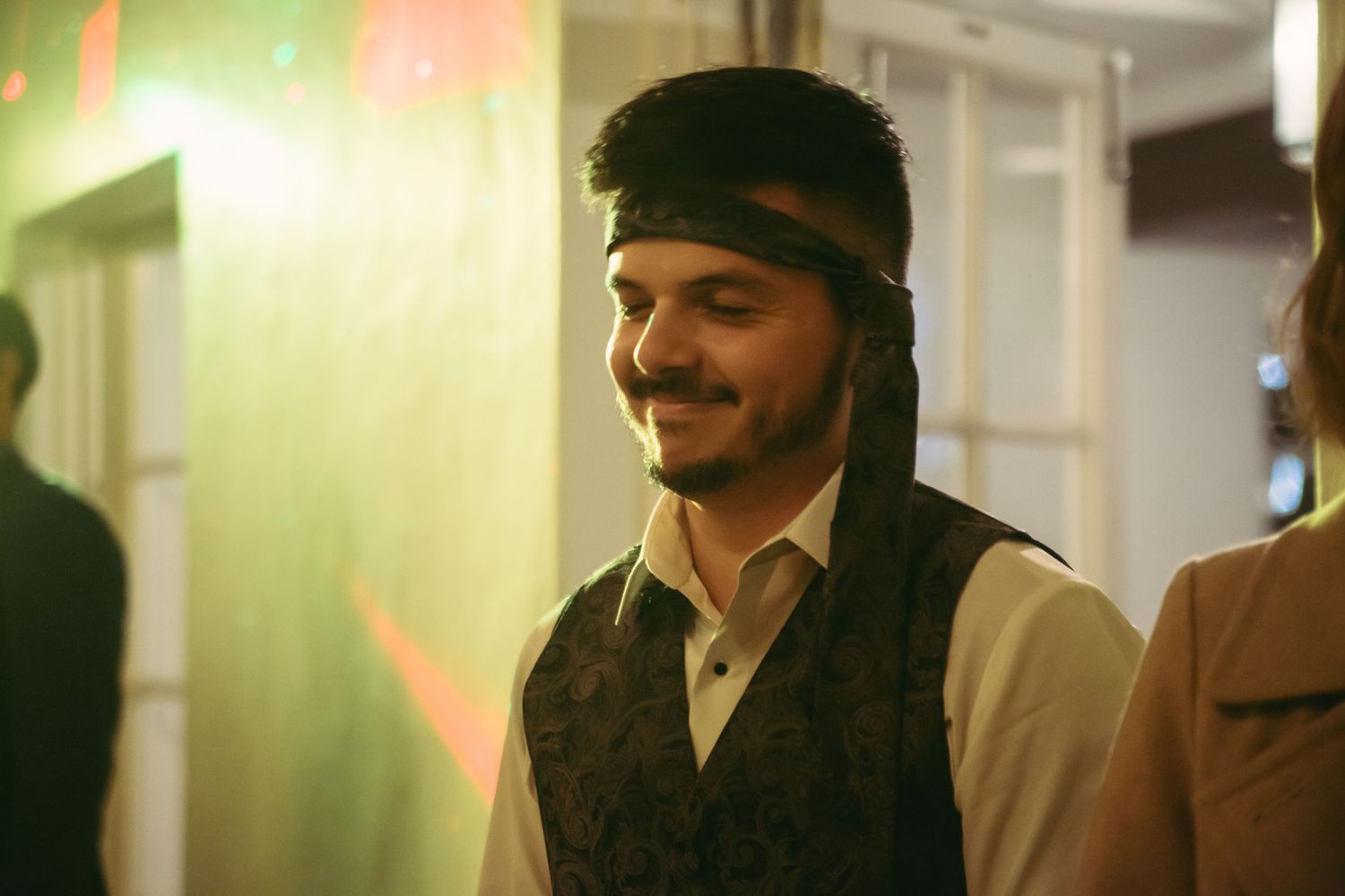 Young man wearing a vest and headband smiling at a wedding reception with green lights.