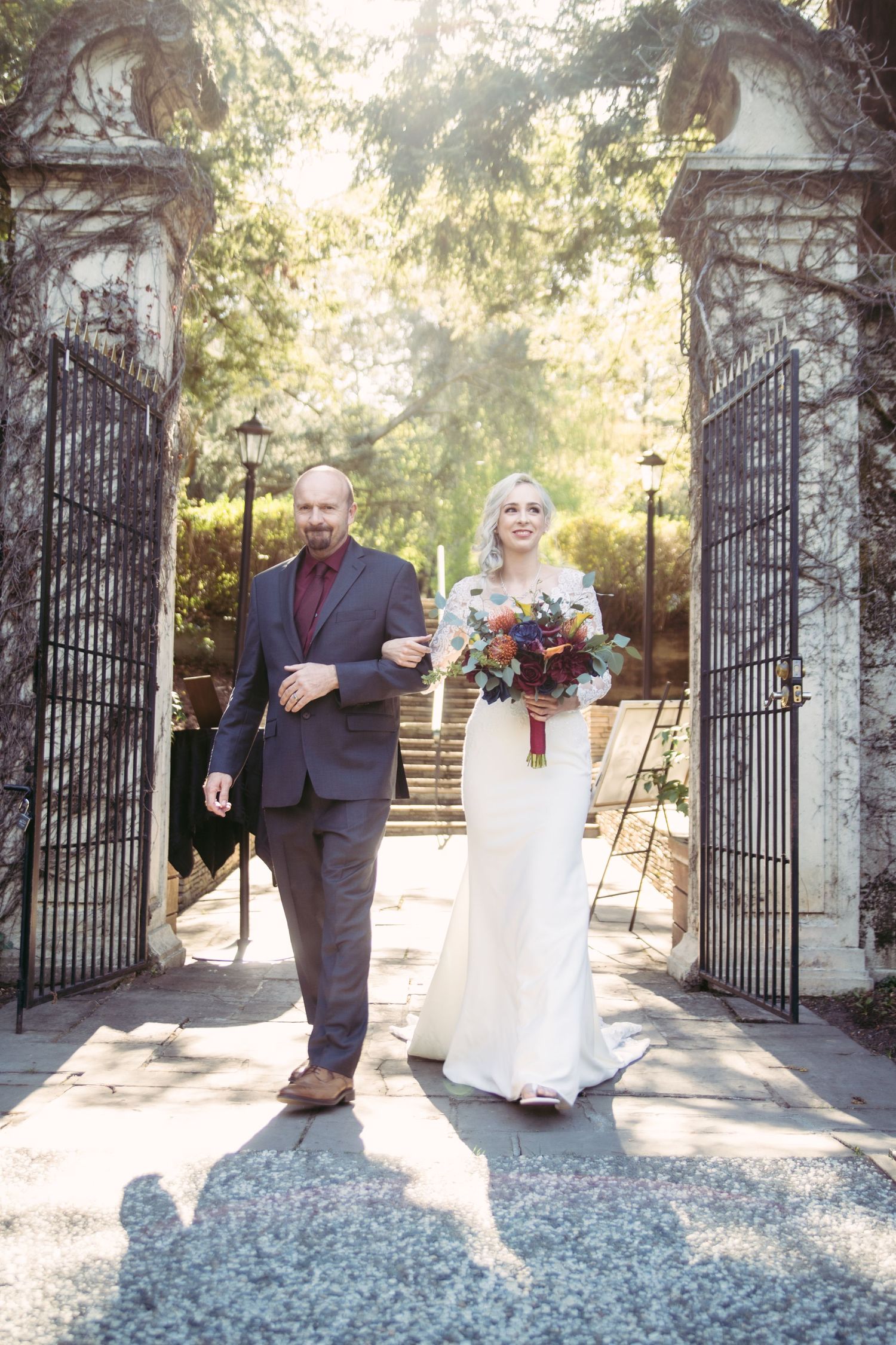 A smiling bride in a white gown holds a vibrant bouquet while walking with her escort through iron garden gates.