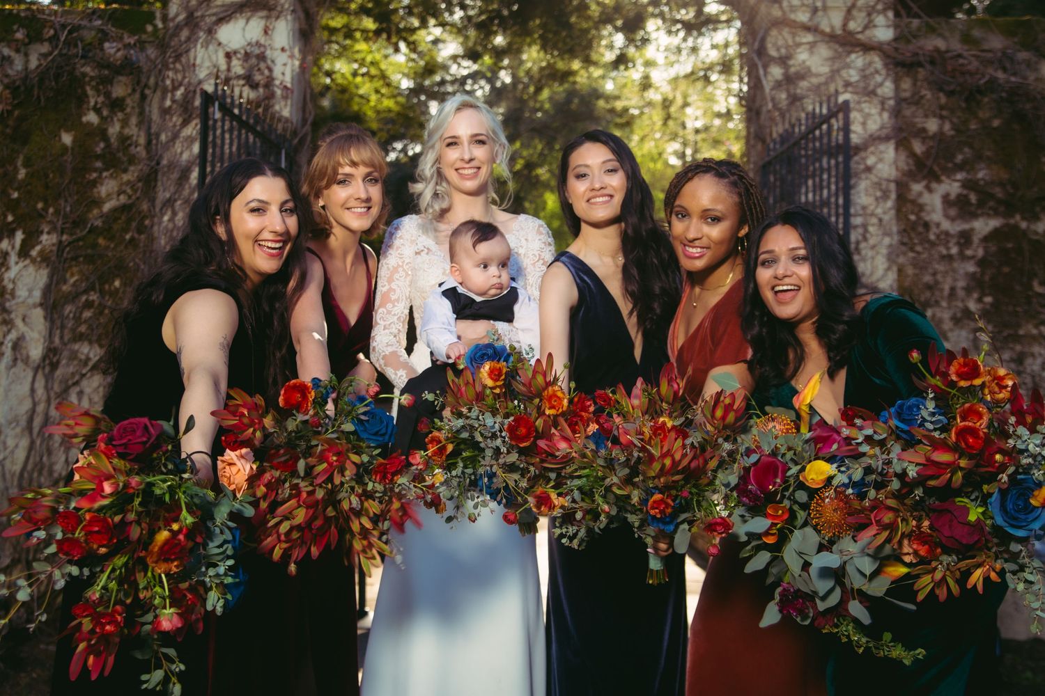 Smiling bride and bridesmaids in dark velvet dresses display lush multicolored bouquets before ivy-covered stone gates.