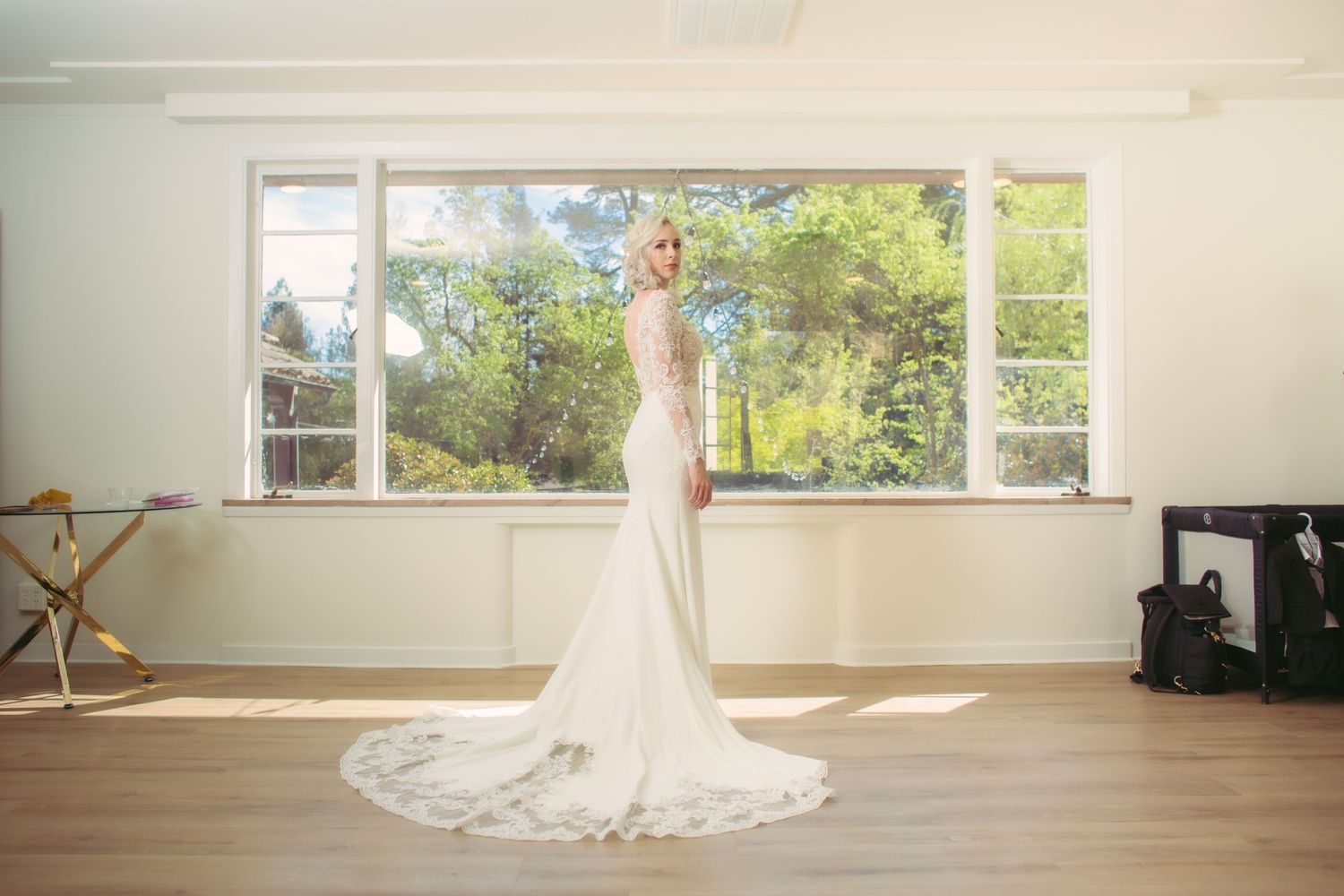 Elegant bride in long-sleeve lace-back wedding dress posing by large sunlit windows with garden view.