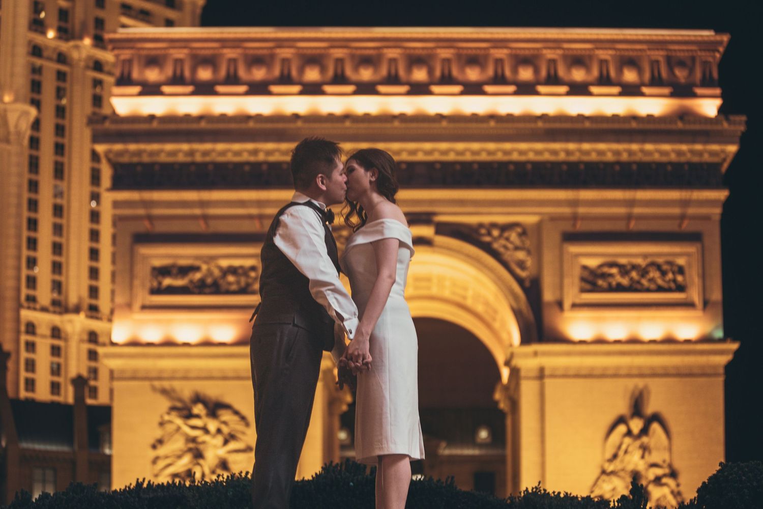 Newlyweds kiss in front of the illuminated Paris Las Vegas Arc de Triomphe replica at night.