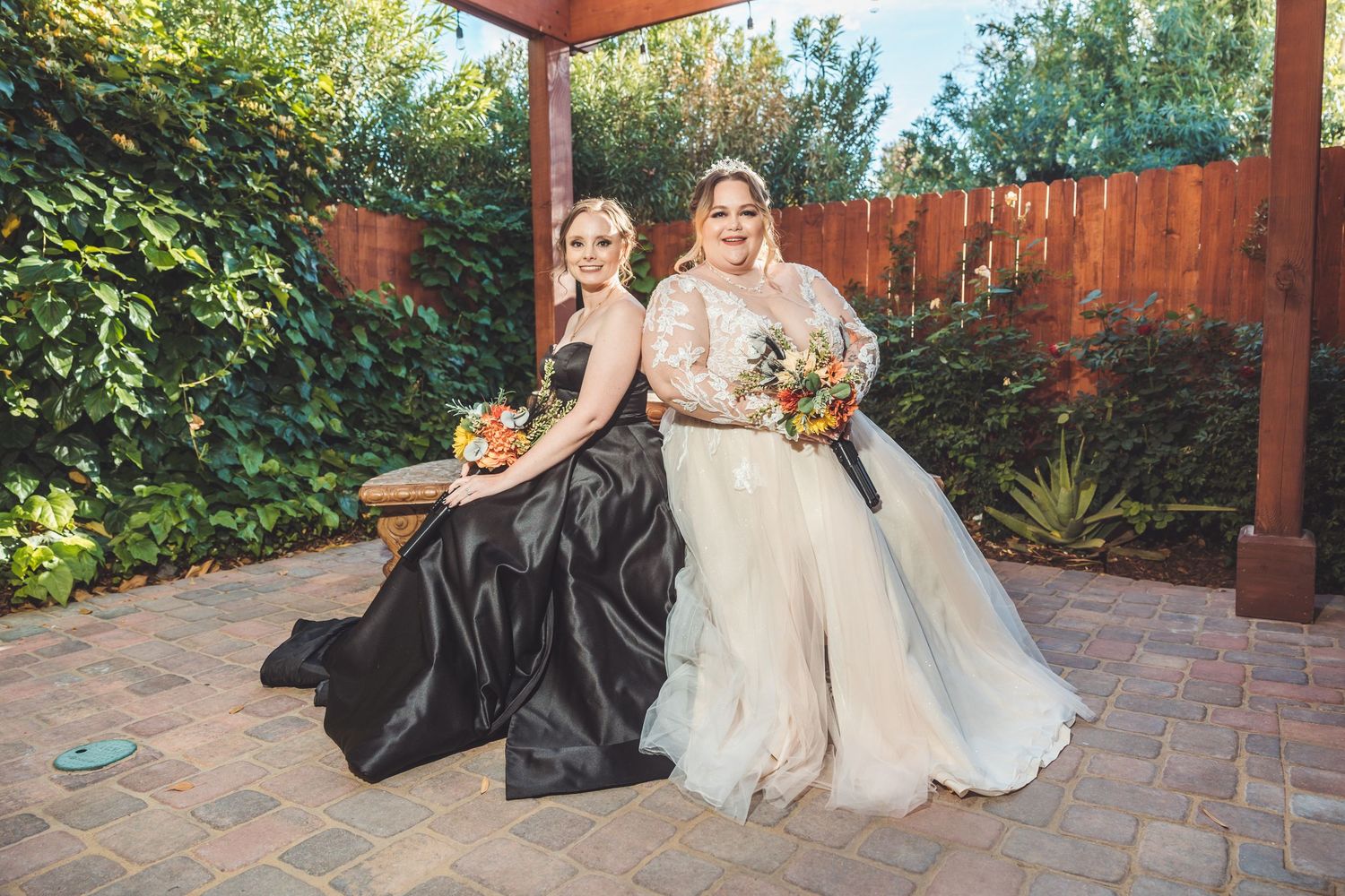 Two brides in white lace and black gowns pose together on brick patio outdoors.