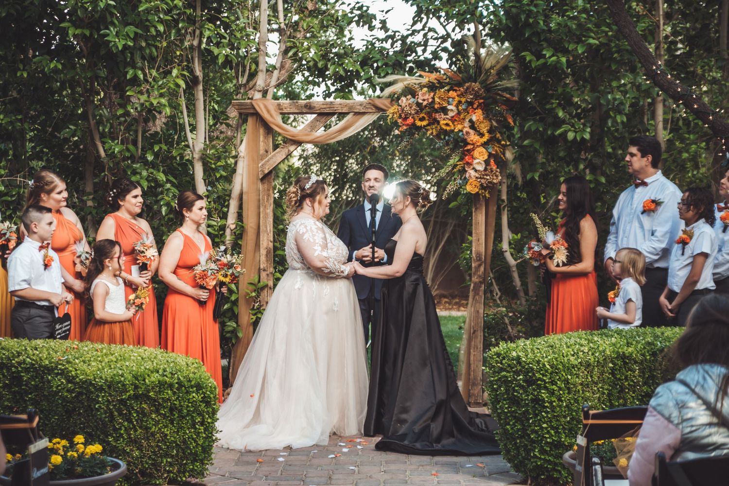 Two brides exchange vows under a rustic wooden arch adorned with fall flowers, surrounded by bridesmaids in orange dresses.