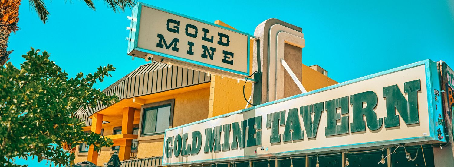 Exterior sign of Gold Mine Tavern bar and restaurant with bright teal lettering on a sunny day.