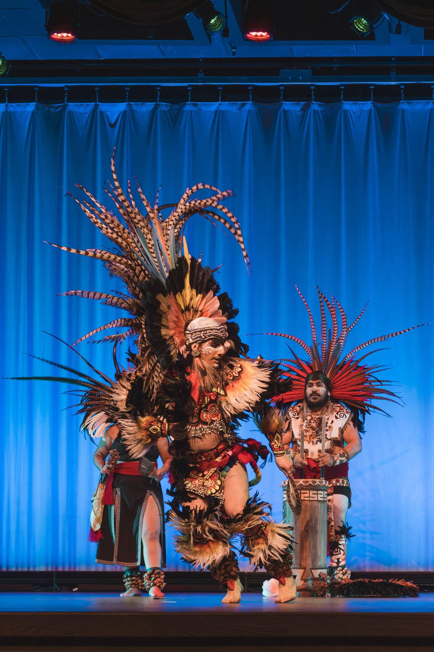 Aztec dancers in traditional feathered headdresses and costumes performing on a blue-lit stage.