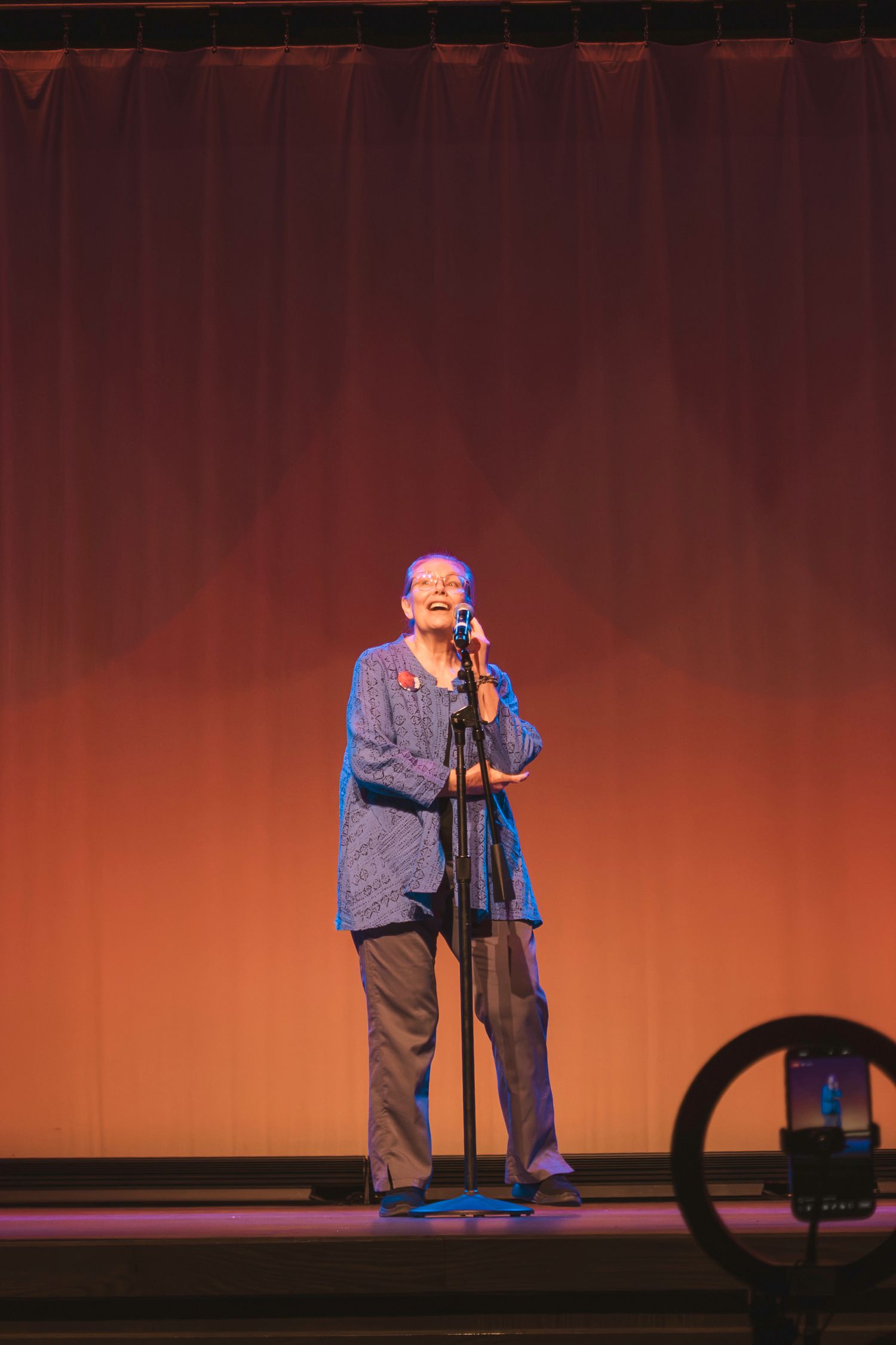 Elderly woman performing on stage at a microphone stand under warm amber stage lighting.