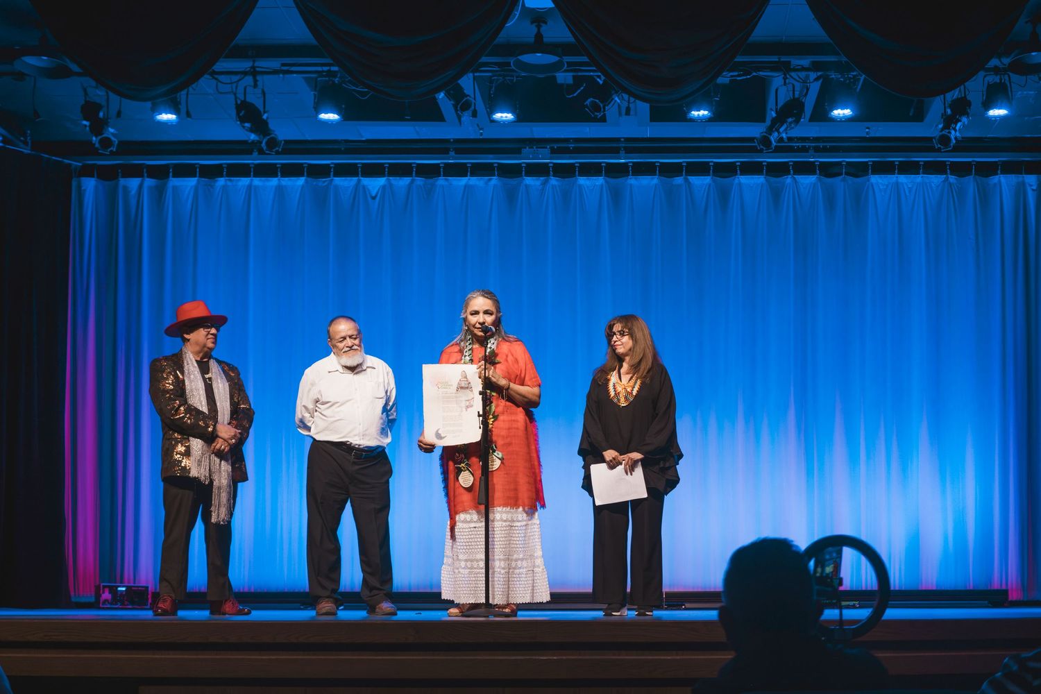 Four people stand on a blue-lit stage, one holding a certificate during a formal presentation ceremony.