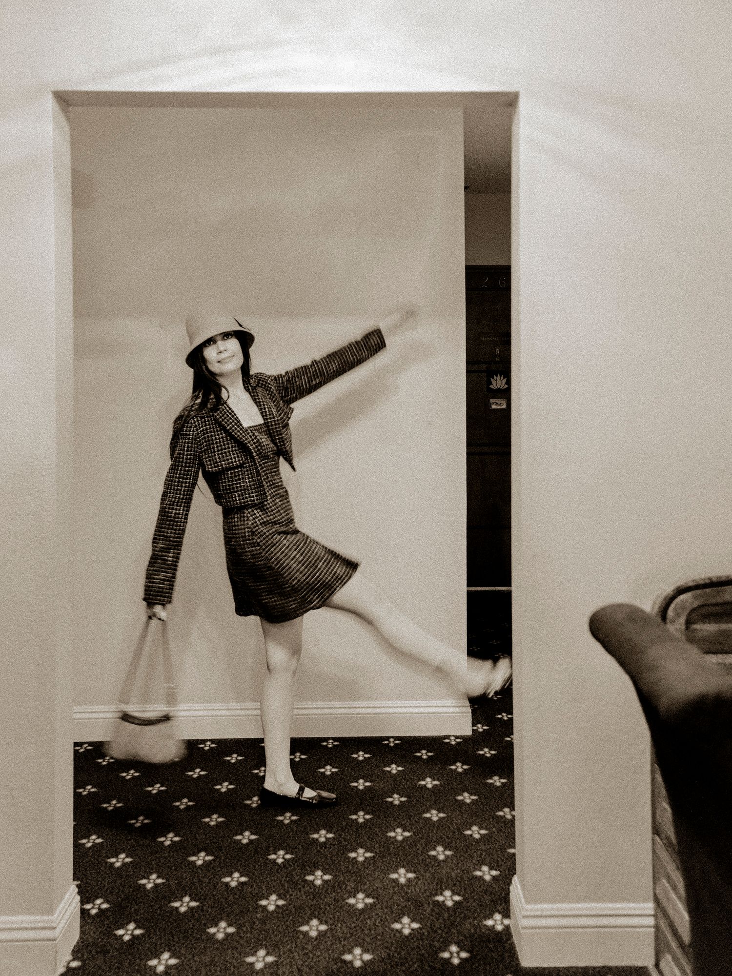 Woman in tweed jacket, mini skirt, and bucket hat poses dynamically in a doorway on a patterned carpet, black and white photo.