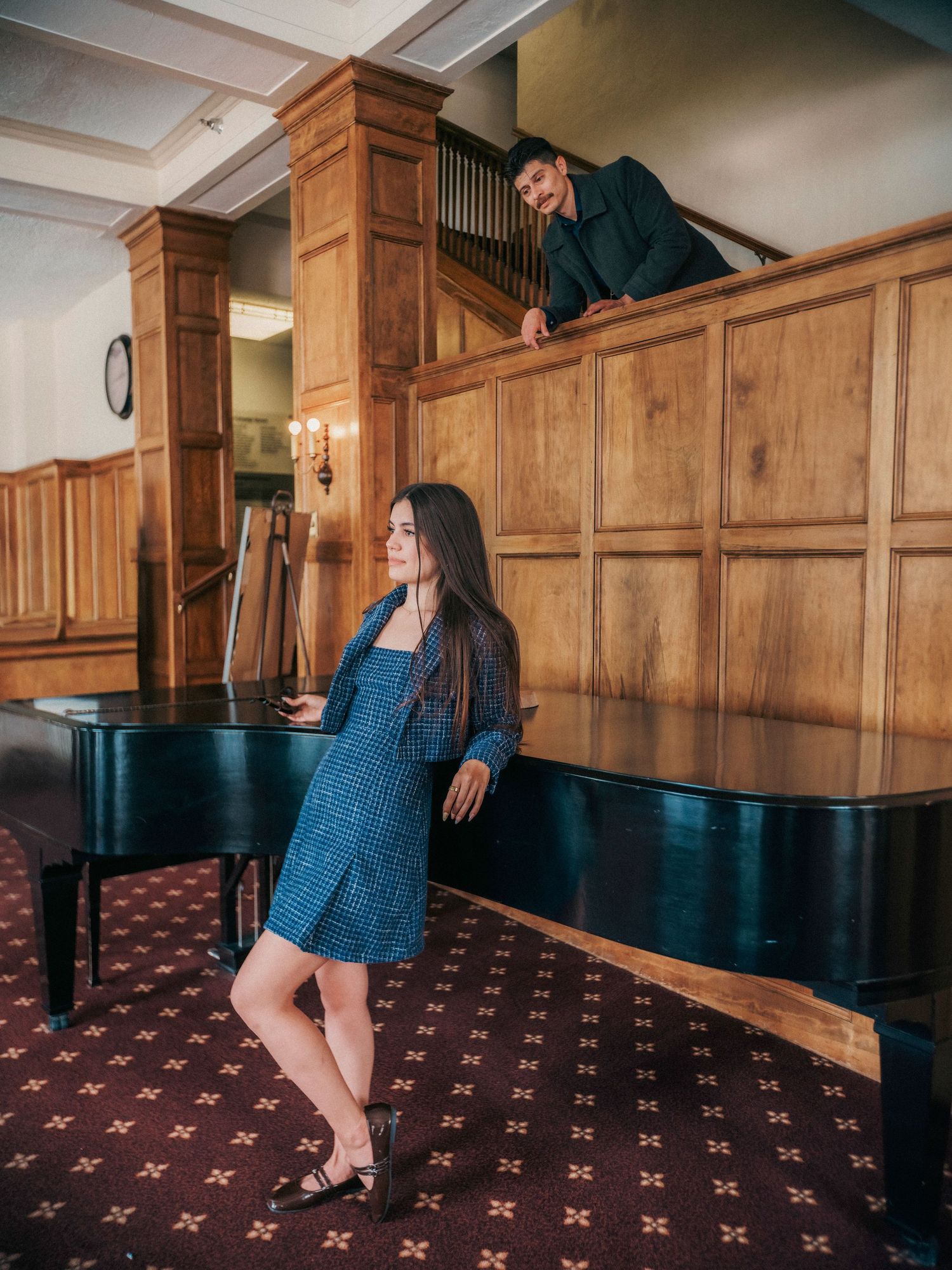 A woman in a blue dress leans on a grand piano while a man watches from a wooden balcony above.