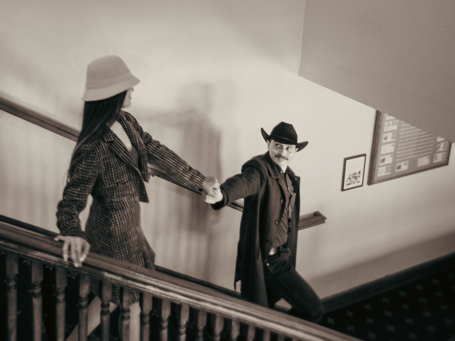 A man in a cowboy hat and woman in a white hat hold hands on a vintage staircase in sepia tones.