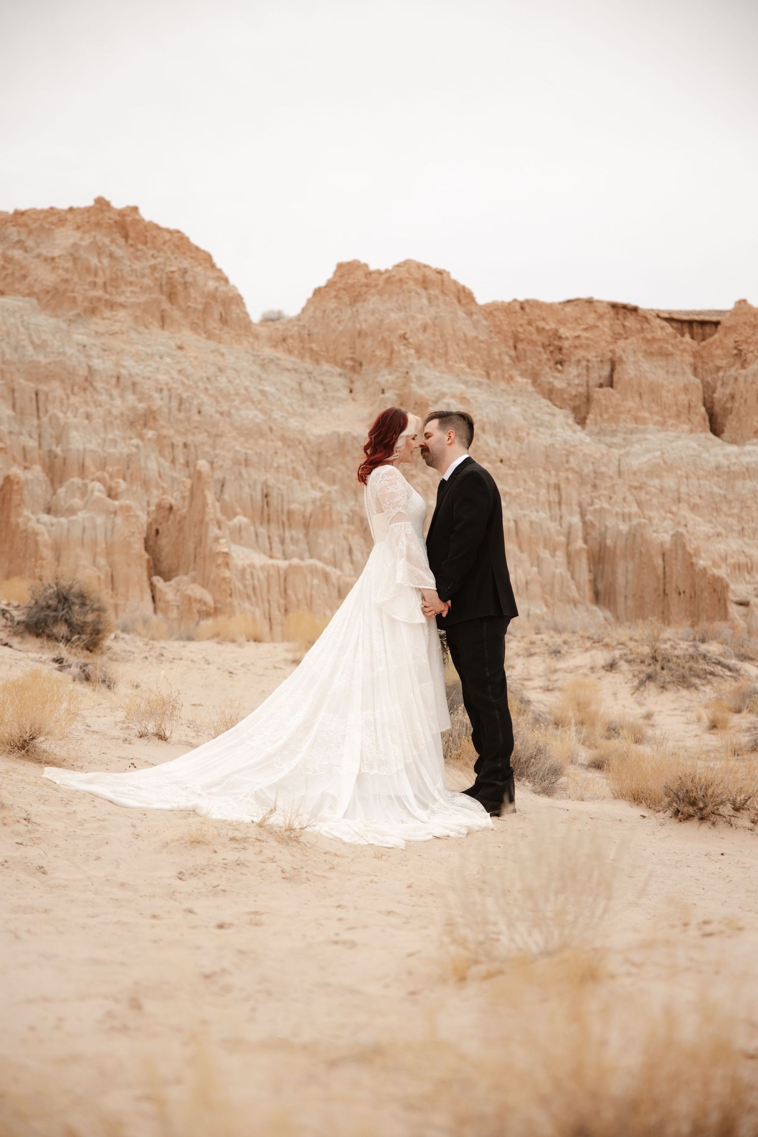 Bride and groom share a romantic moment surrounded by sandy desert landscape and red rock formations.