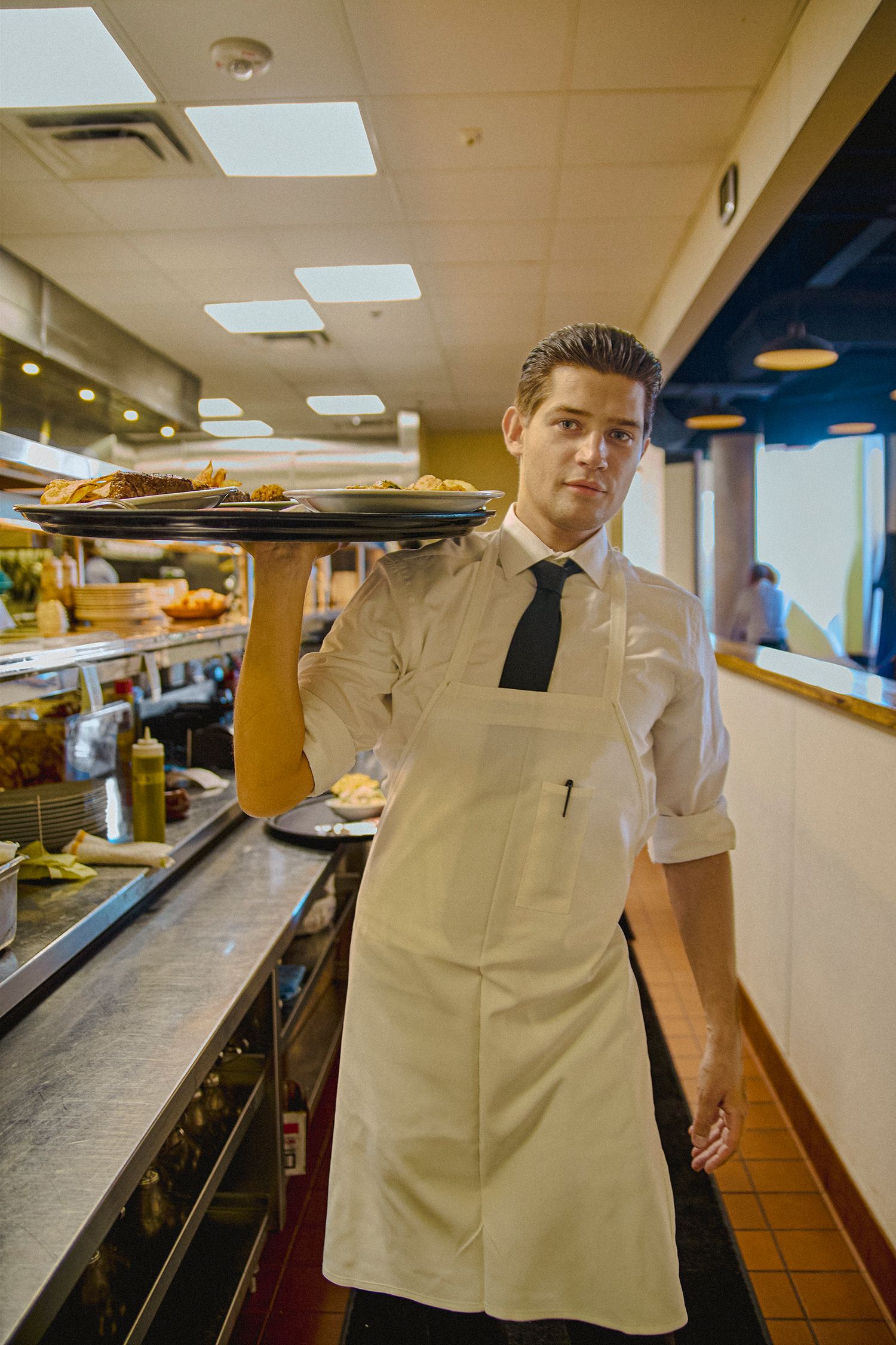 A young waiter in white shirt, tie, and apron carries a food tray in a restaurant kitchen.