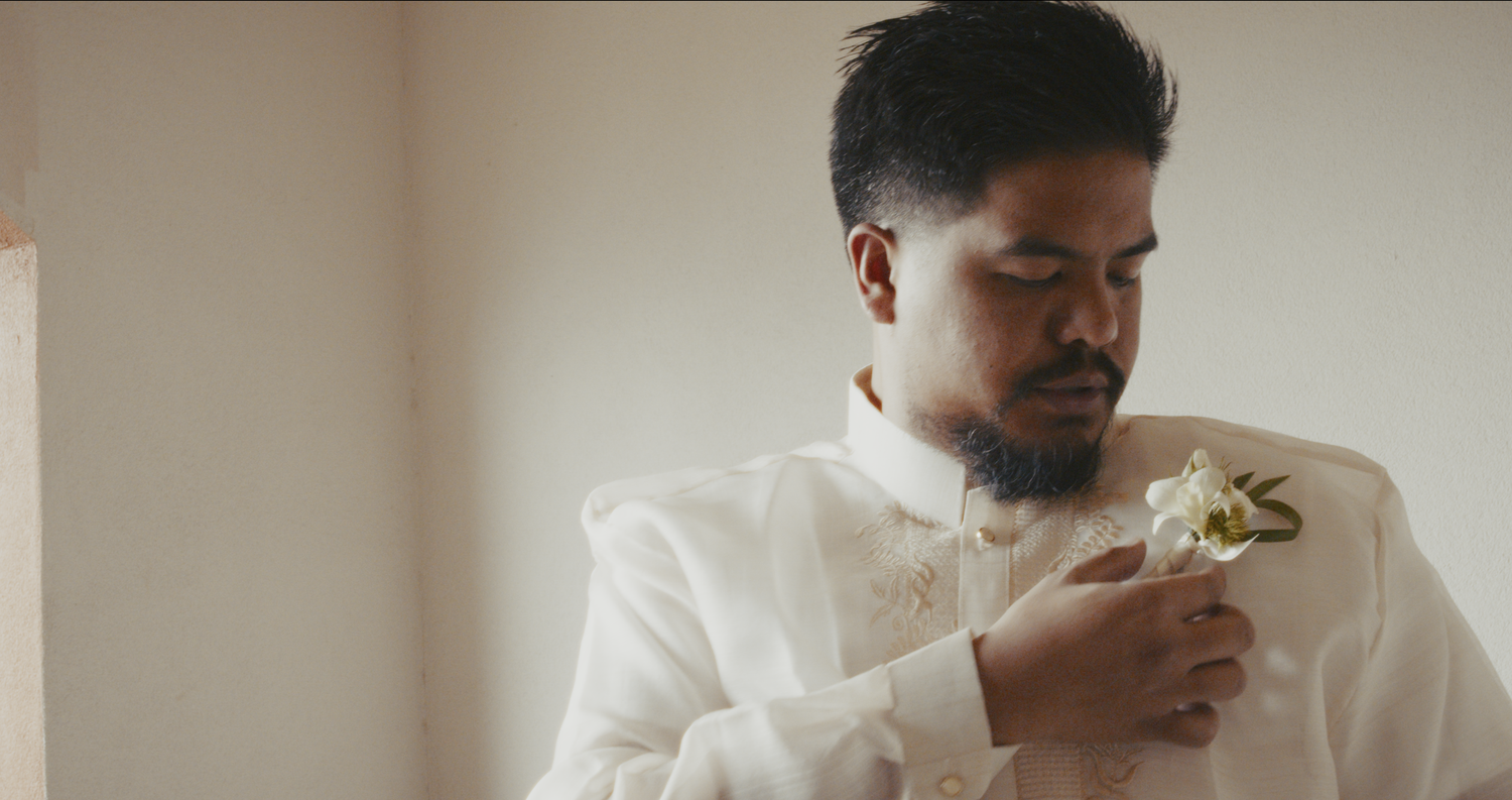 A groom in a white barong looks down while pinning a white floral boutonniere to his lapel.