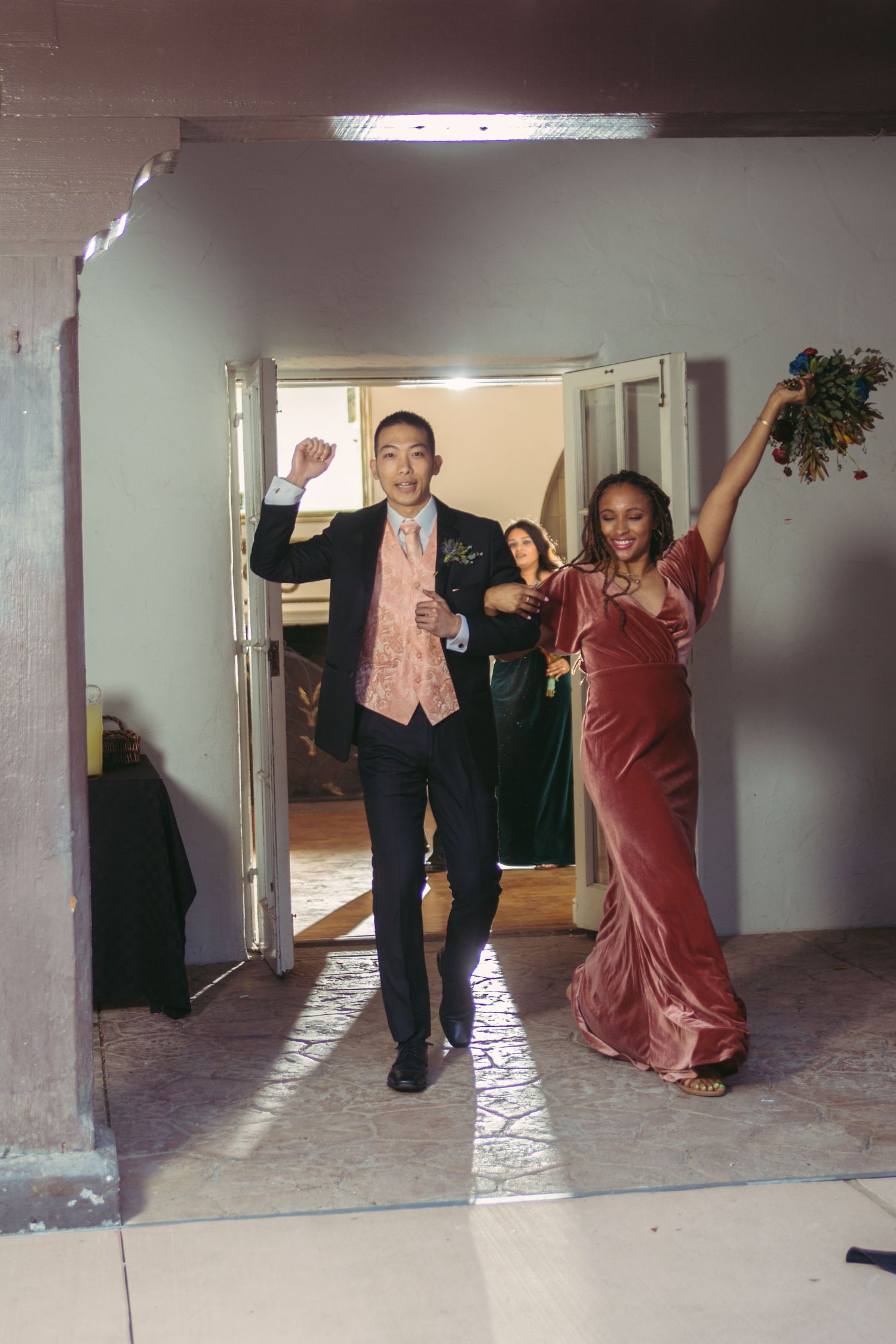 Couple in pink velvet dress and navy suit joyfully entering a wedding reception holding a festive bouquet.