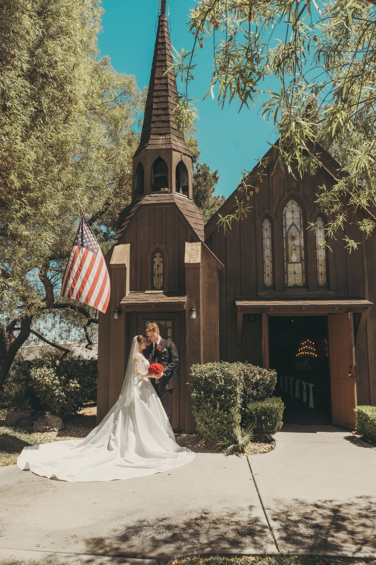 Bride and groom kissing outside a rustic brown wooden chapel with a steeple and American flag on a sunny day.