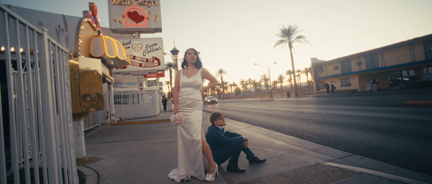 Bride in white gown with groom in blue suit on Las Vegas Strip sidewalk near iconic neon signs at sunset.