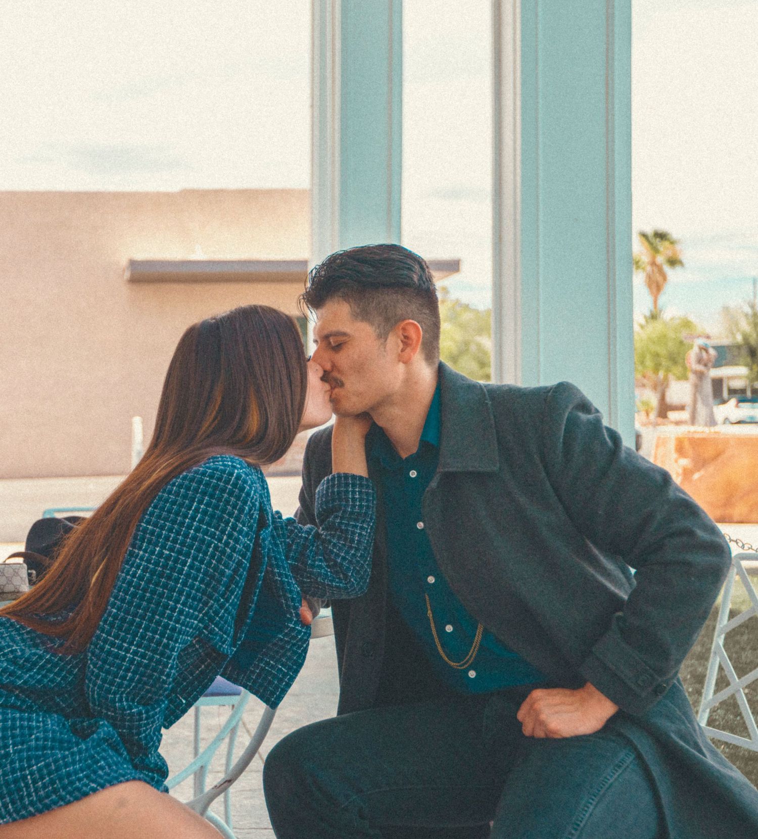 A couple shares a romantic kiss at an outdoor cafe, dressed in coordinating teal outfits on a sunny day.