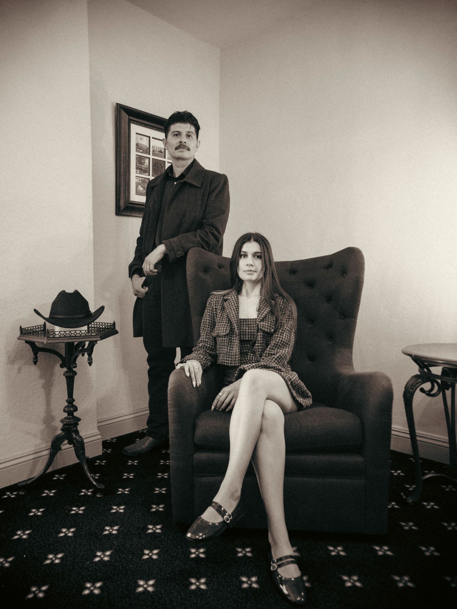 Man in black coat stands beside woman seated in tufted armchair in moody black and white portrait.