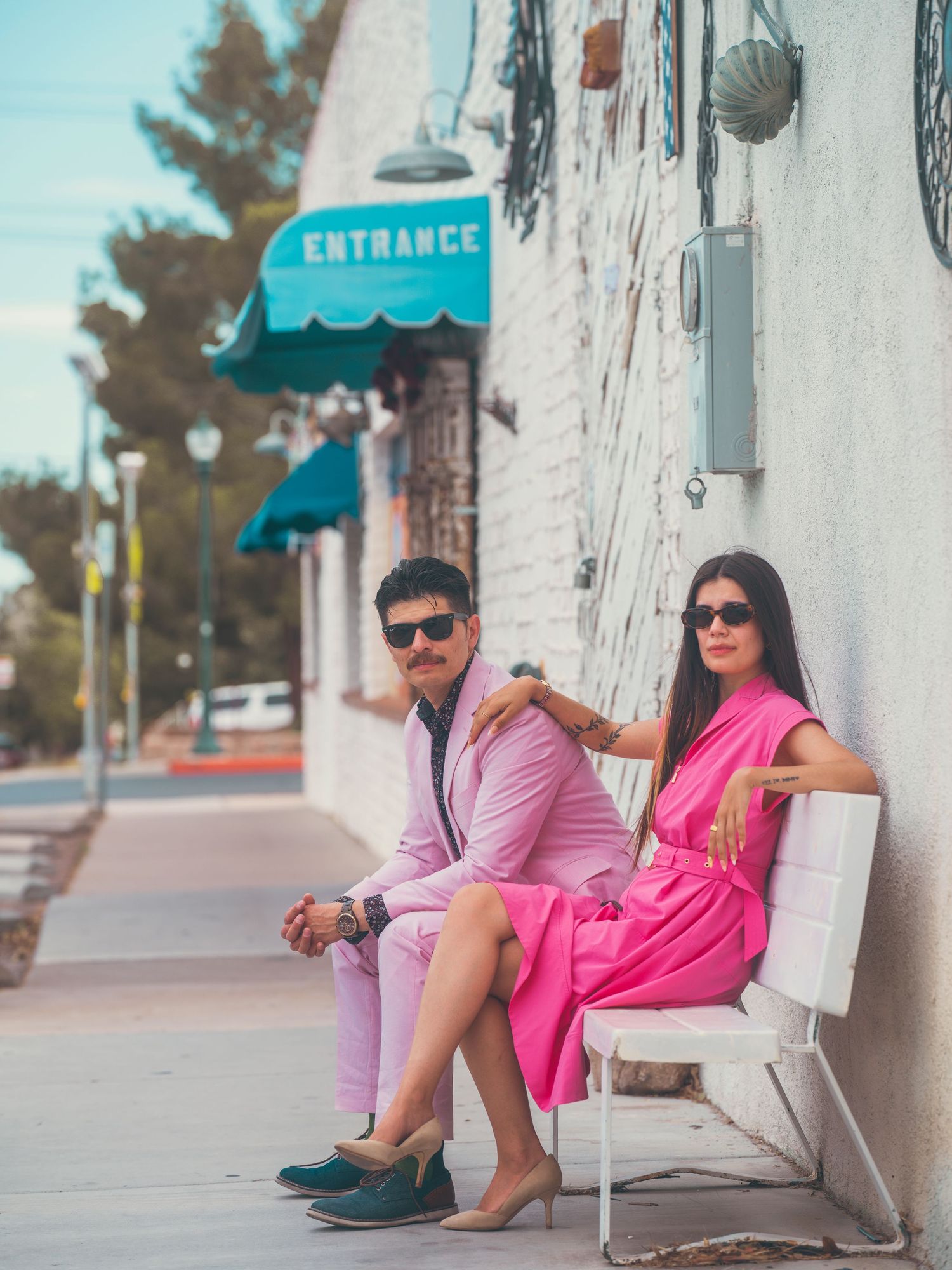 A stylish couple in matching pink outfits sitting on a white bench outside a building with an Entrance sign.