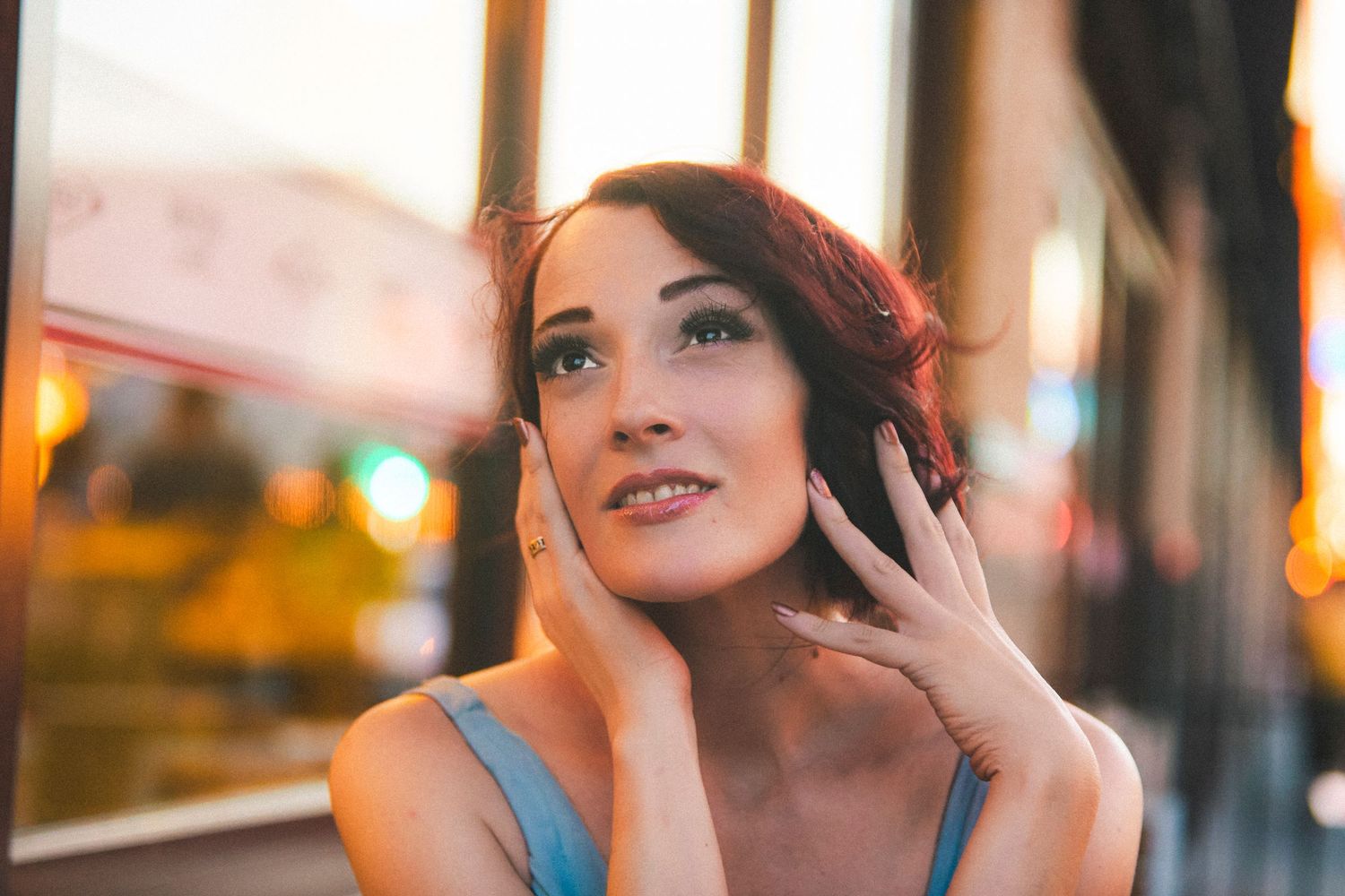 Young woman with dark hair poses thoughtfully outdoors at sunset, gently touching her neck with a soft smile.