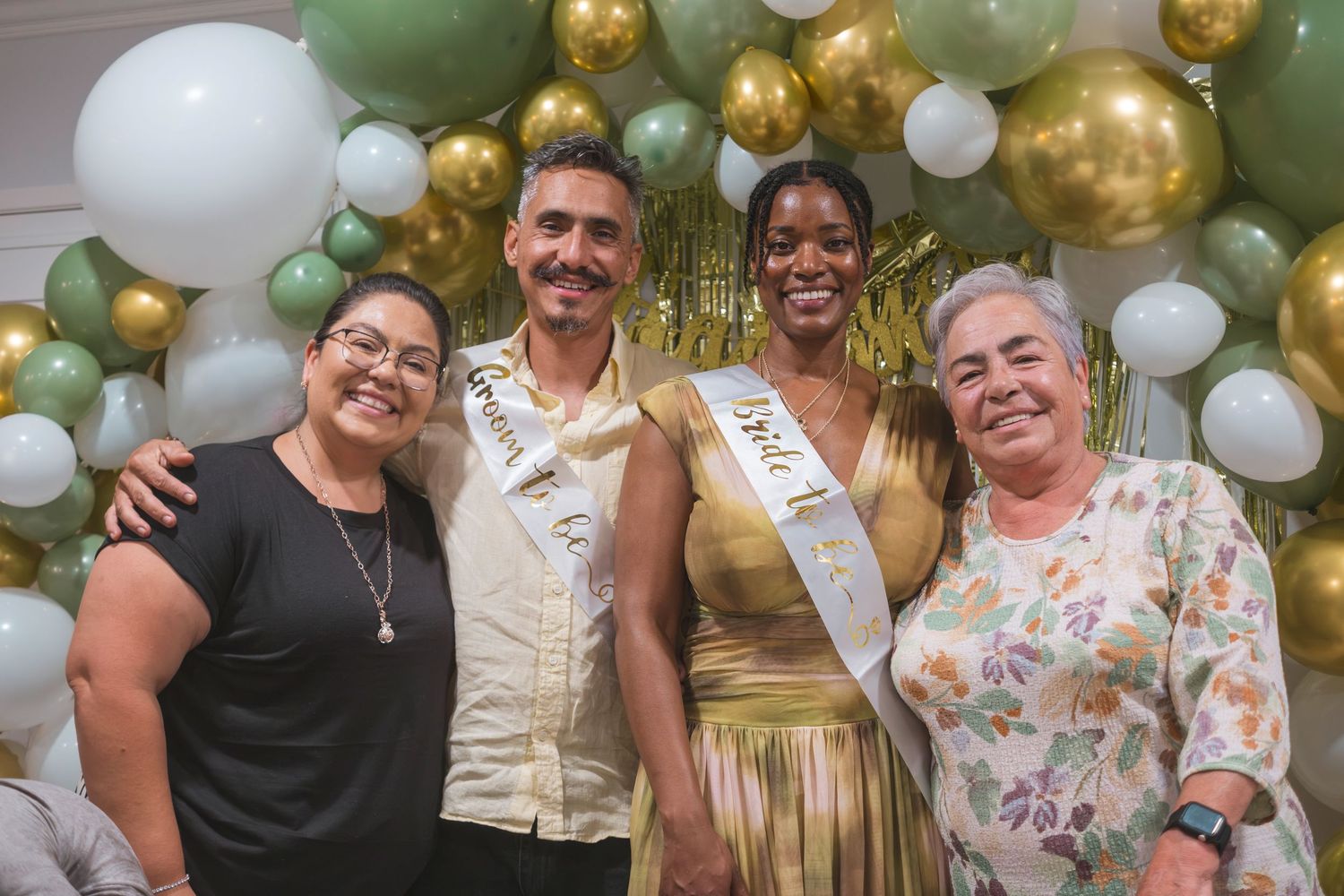 Four people posing together at a festive event with green, white, and gold balloon decorations.