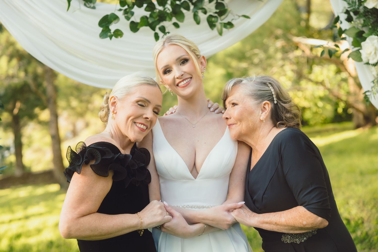 Bride in white gown flanked by two women in black dresses under floral arch.