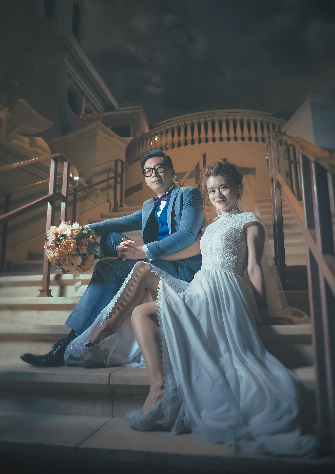 Couple in wedding attire sitting on elegant staircase, groom in blue suit holding bouquet, bride in white lace gown.