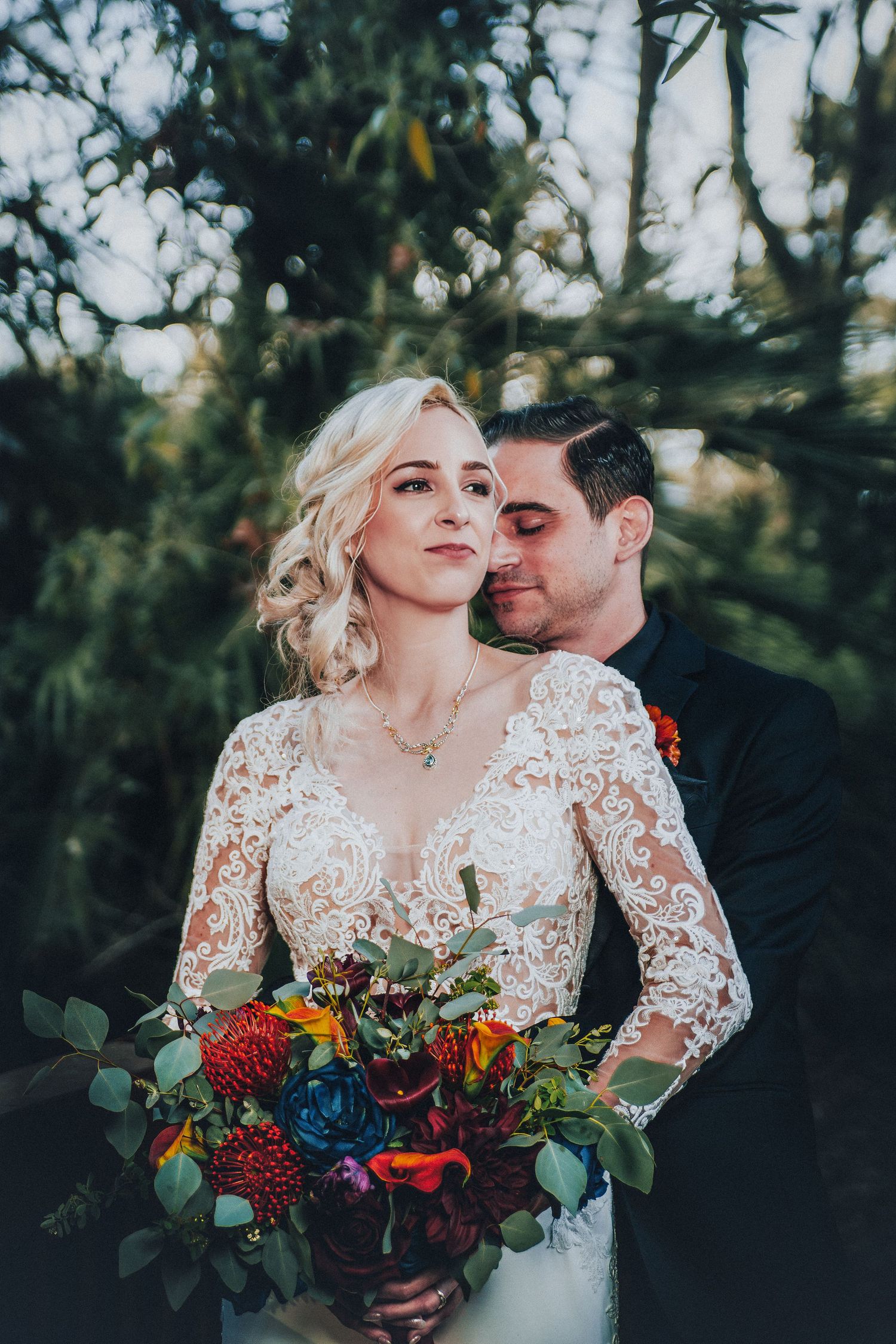 Bride in lace gown holds colorful bouquet as groom in black suit embraces her outdoors among lush green trees.