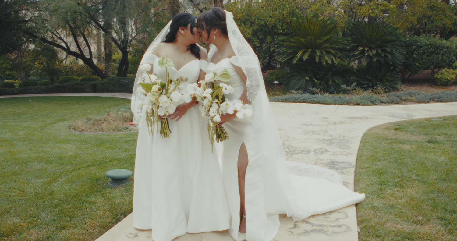 Two brides in white gowns holding floral bouquets share an intimate moment in a garden pathway.