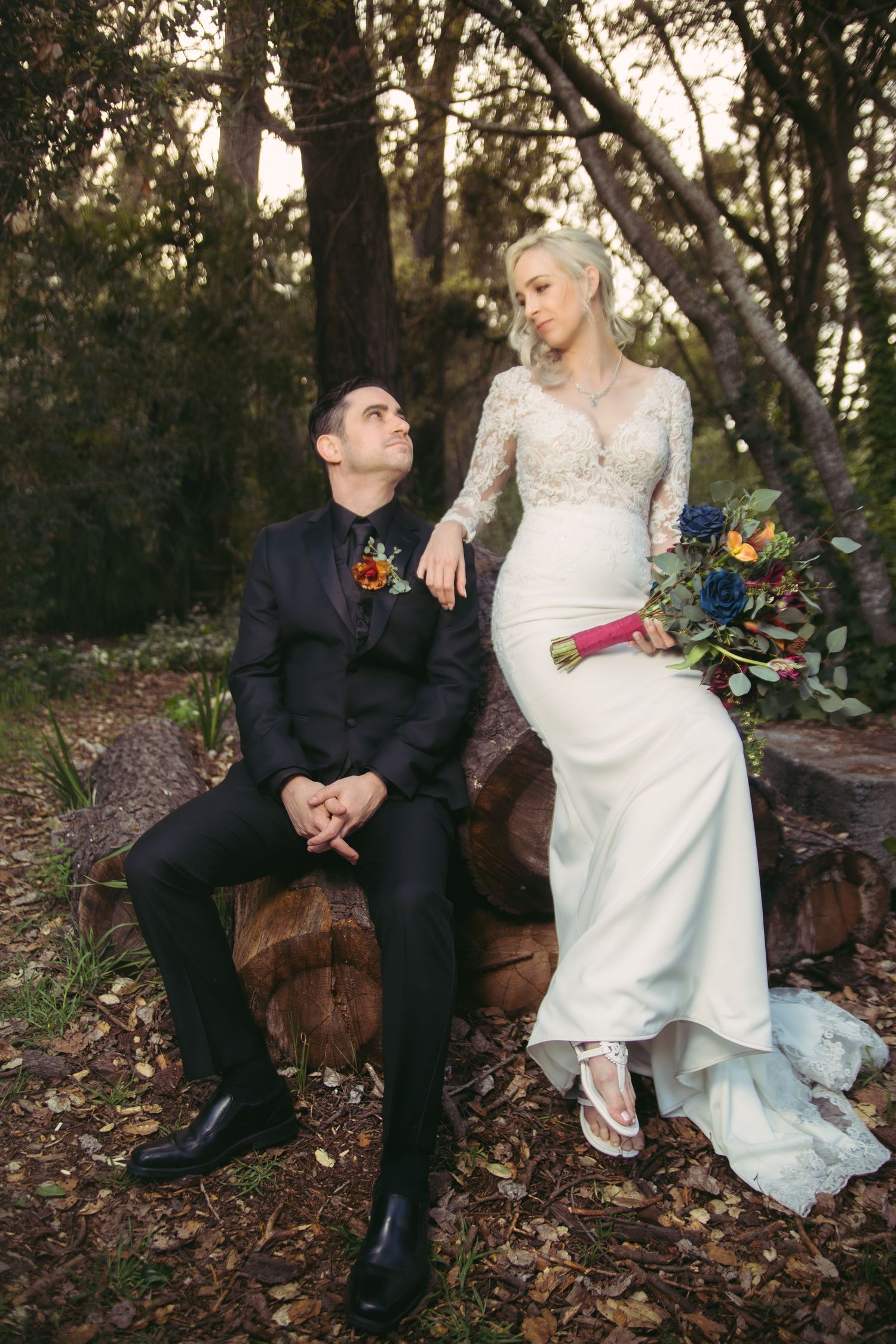 Groom gazes up at smiling bride seated on logs in forest, bride holds colorful floral bouquet in lace gown.