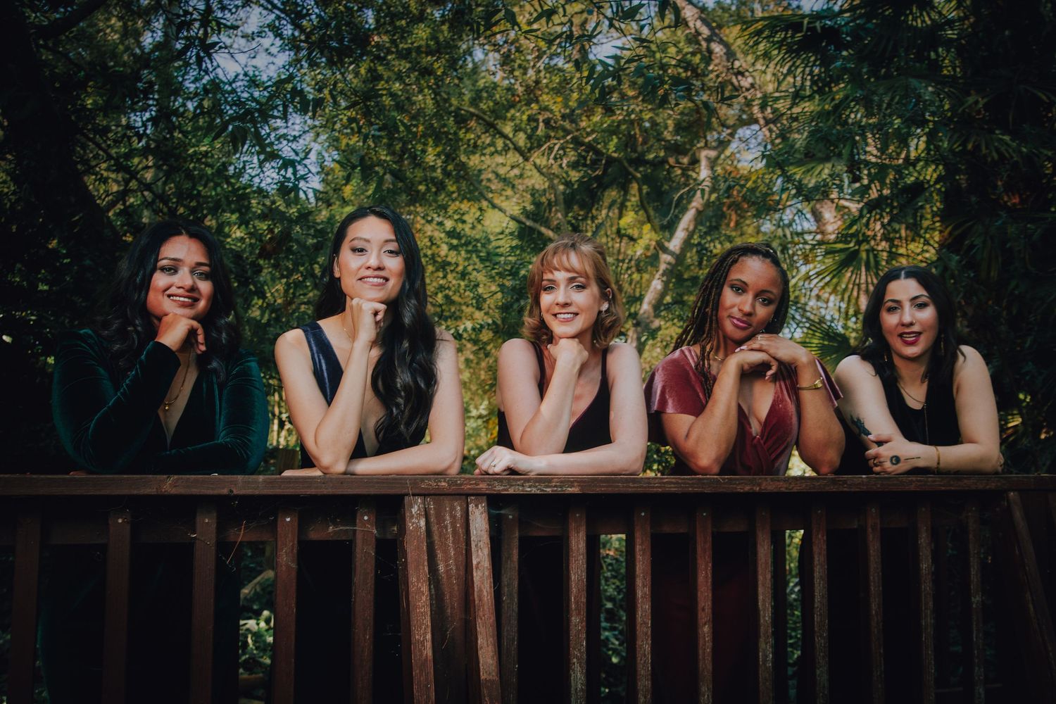 Five young women smiling and leaning on a wooden railing outdoors, surrounded by lush green trees.