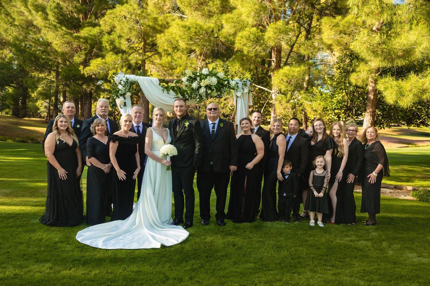 Wedding party in black attire poses with bride and groom outdoors under a floral arch in a park.