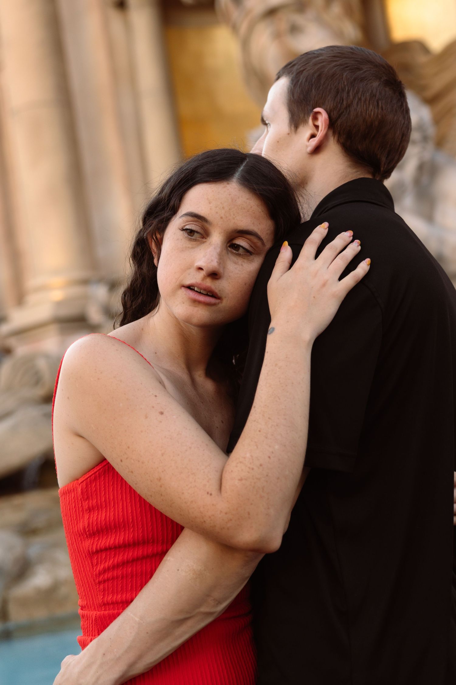 Close portrait of woman in red dress with freckles embracing man in black near stone architecture.