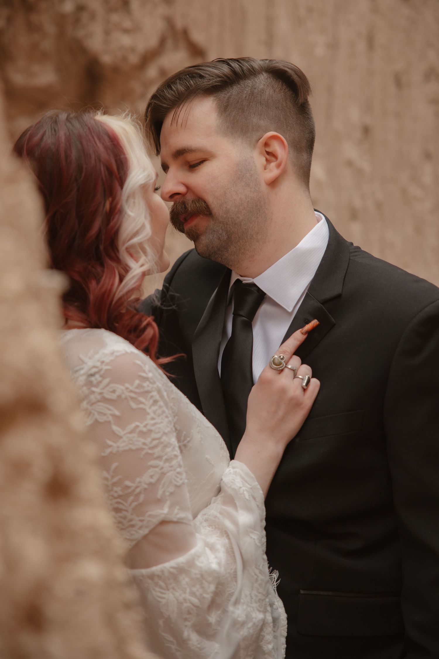 Groom kisses bride wearing lace wedding dress while she holds his lapel with jeweled rings.