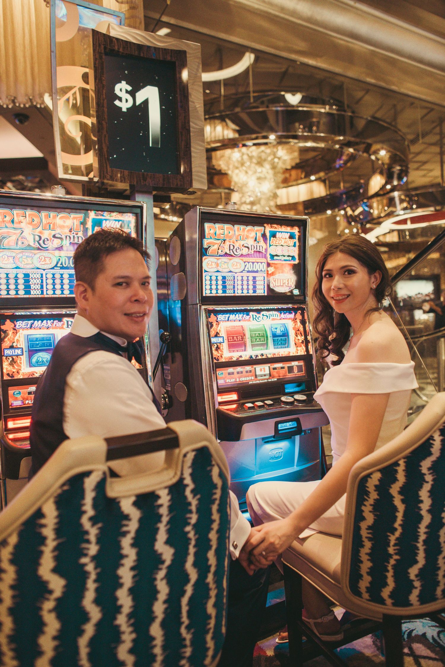 Newlyweds sitting at slot machines in Las Vegas casino, smiling and holding hands in wedding attire.