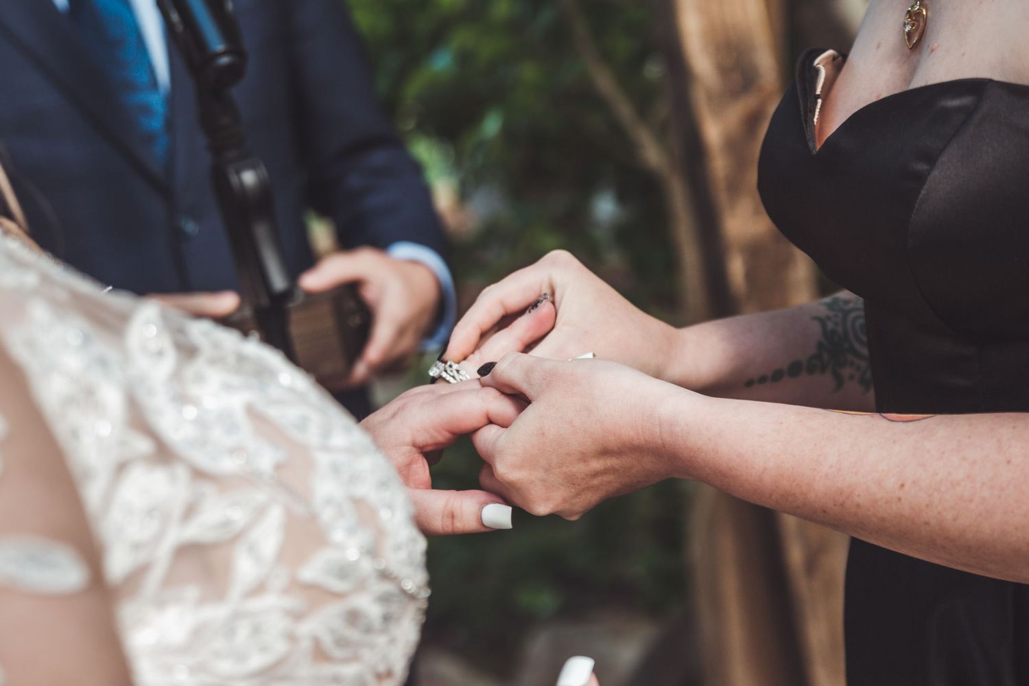 Close-up of a wedding ring exchange ceremony, bride in lace dress receiving ring from officiant outdoors.