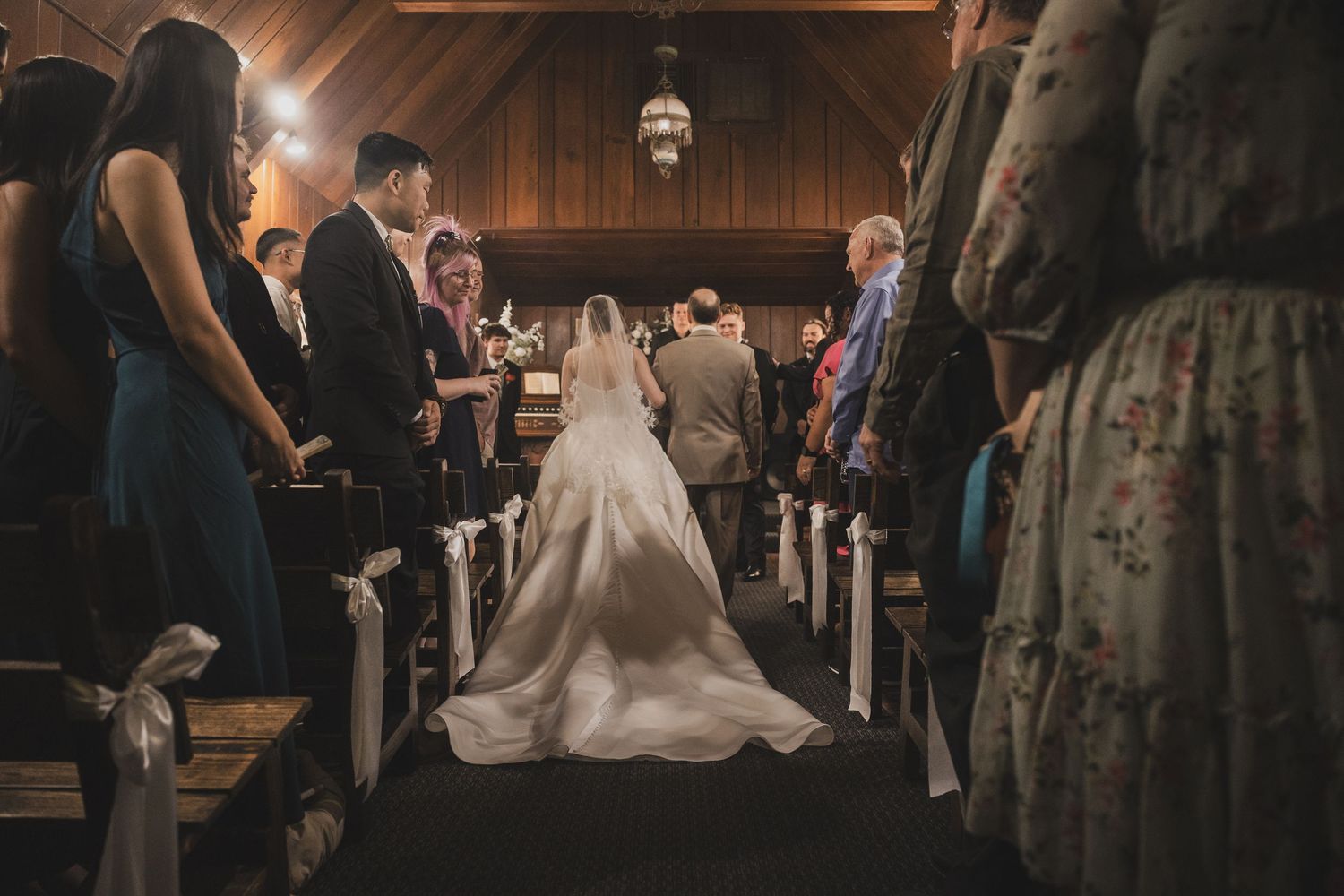 Bride walking down the aisle in a white gown at a wooden chapel wedding ceremony with guests seated in pews.