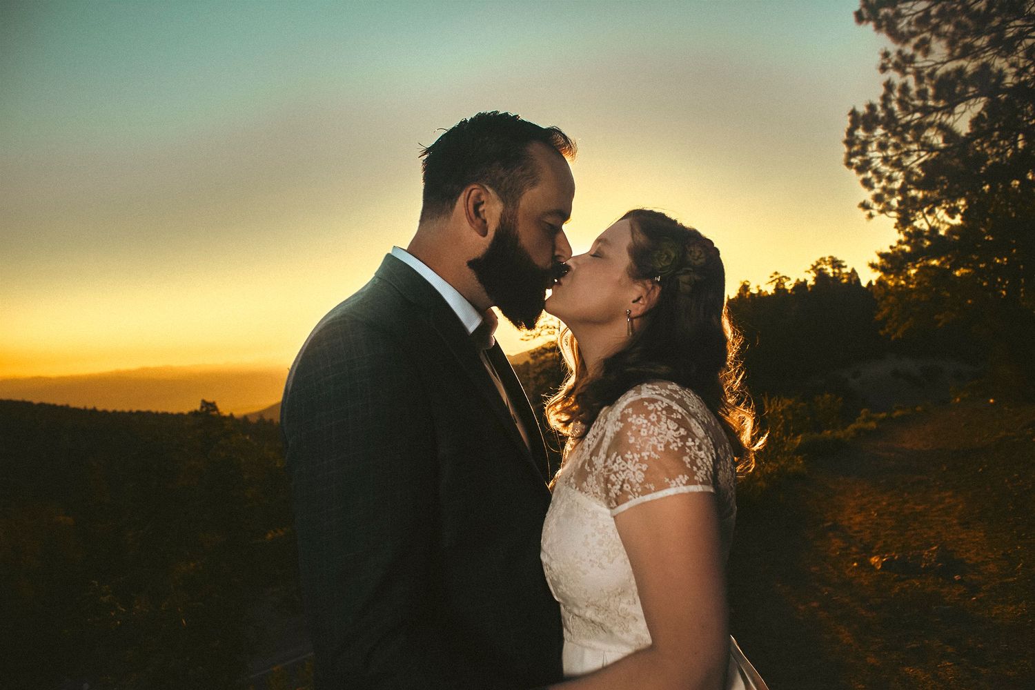 Bride and groom share a romantic sunset kiss outdoors with golden sky backdrop on their wedding day.