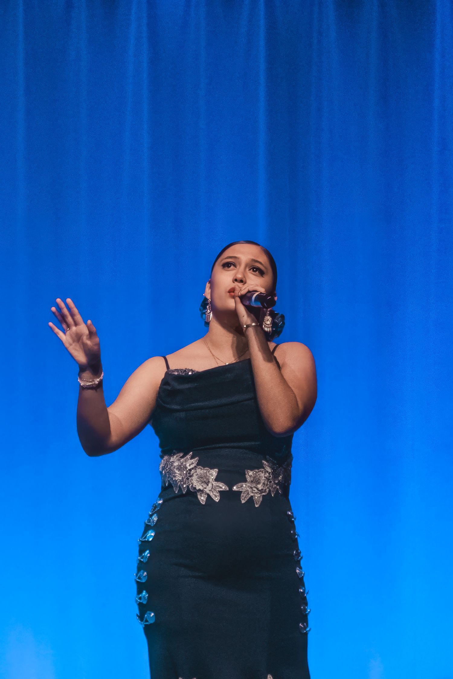 Woman in black embellished dress singing on stage with blue curtain backdrop.