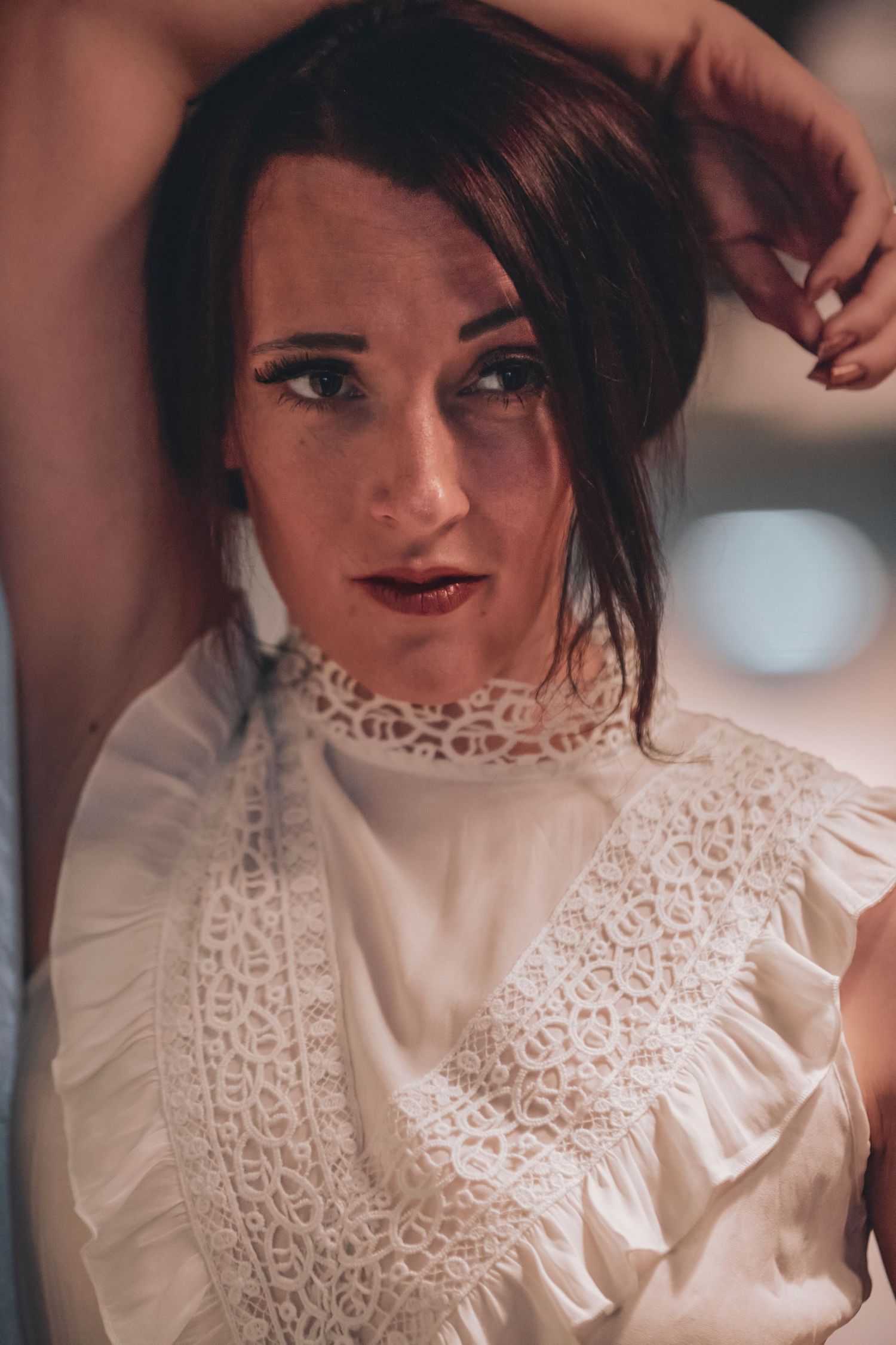 A young woman in a white lace Victorian blouse poses with arms raised, wearing dark lipstick and looking at camera.