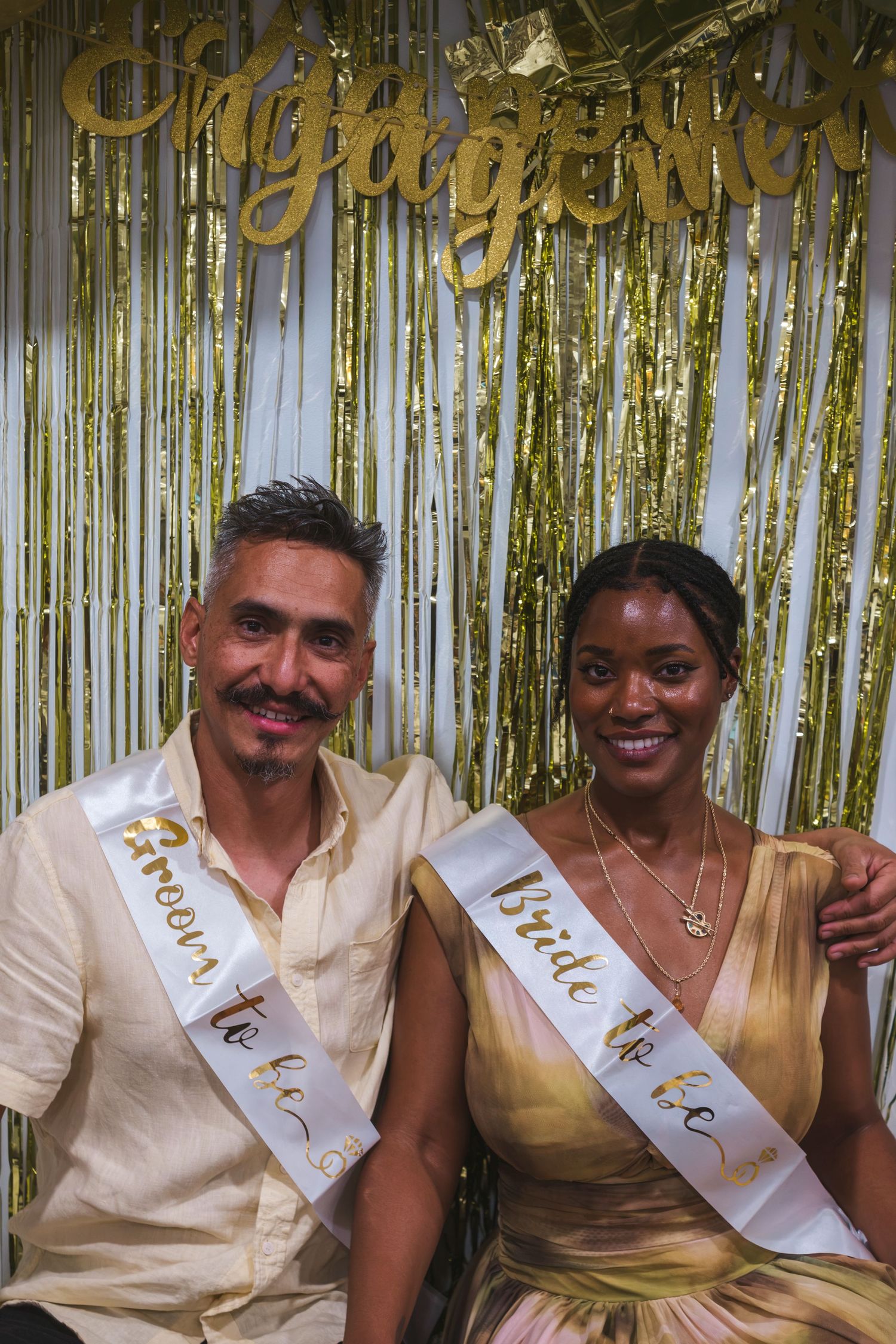 Couple wearing bride and groom-to-be sashes posing at an engagement party with gold tinsel backdrop.