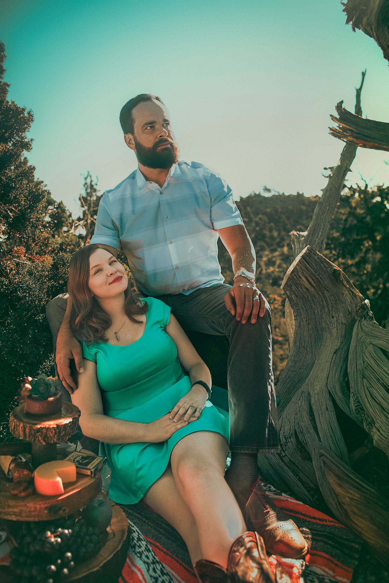 Couple posing outdoors, man with beard in striped shirt sits on log, woman in green dress leans against him.
