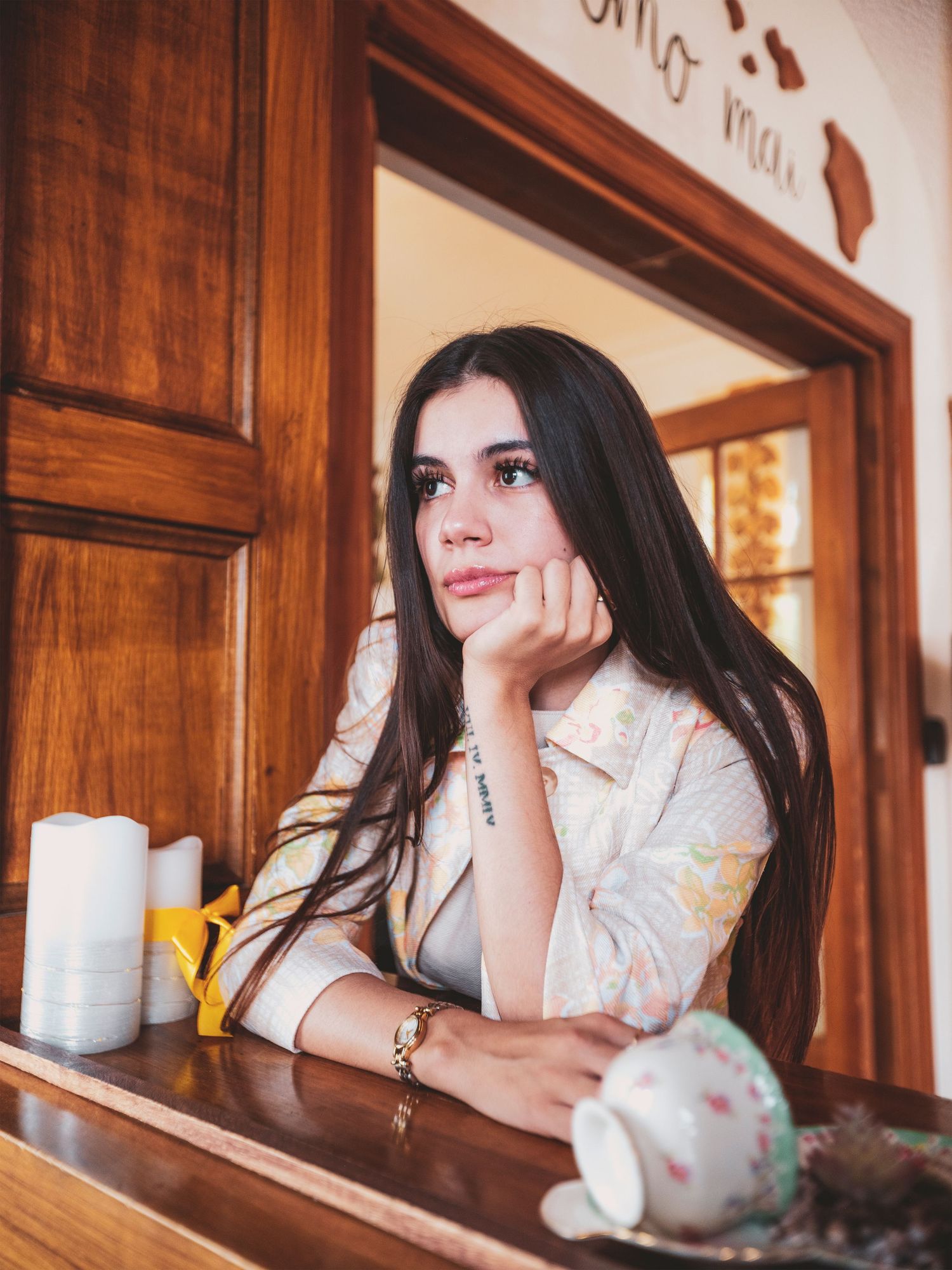 Young woman with long dark hair resting chin on hand at a wooden cafe counter with a coffee cup.