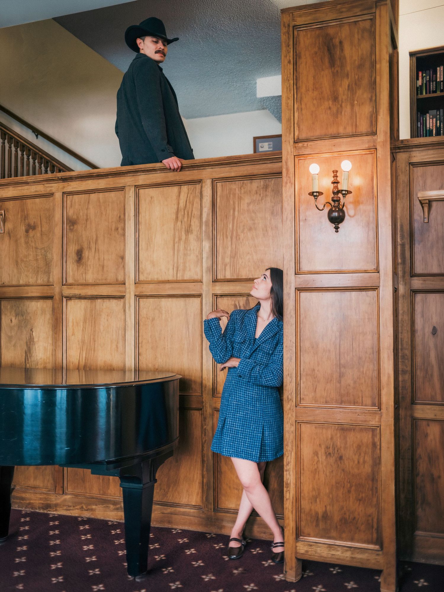 Woman in blue dress looks up at man in black cowboy hat standing on staircase above her in elegant wood-paneled room.