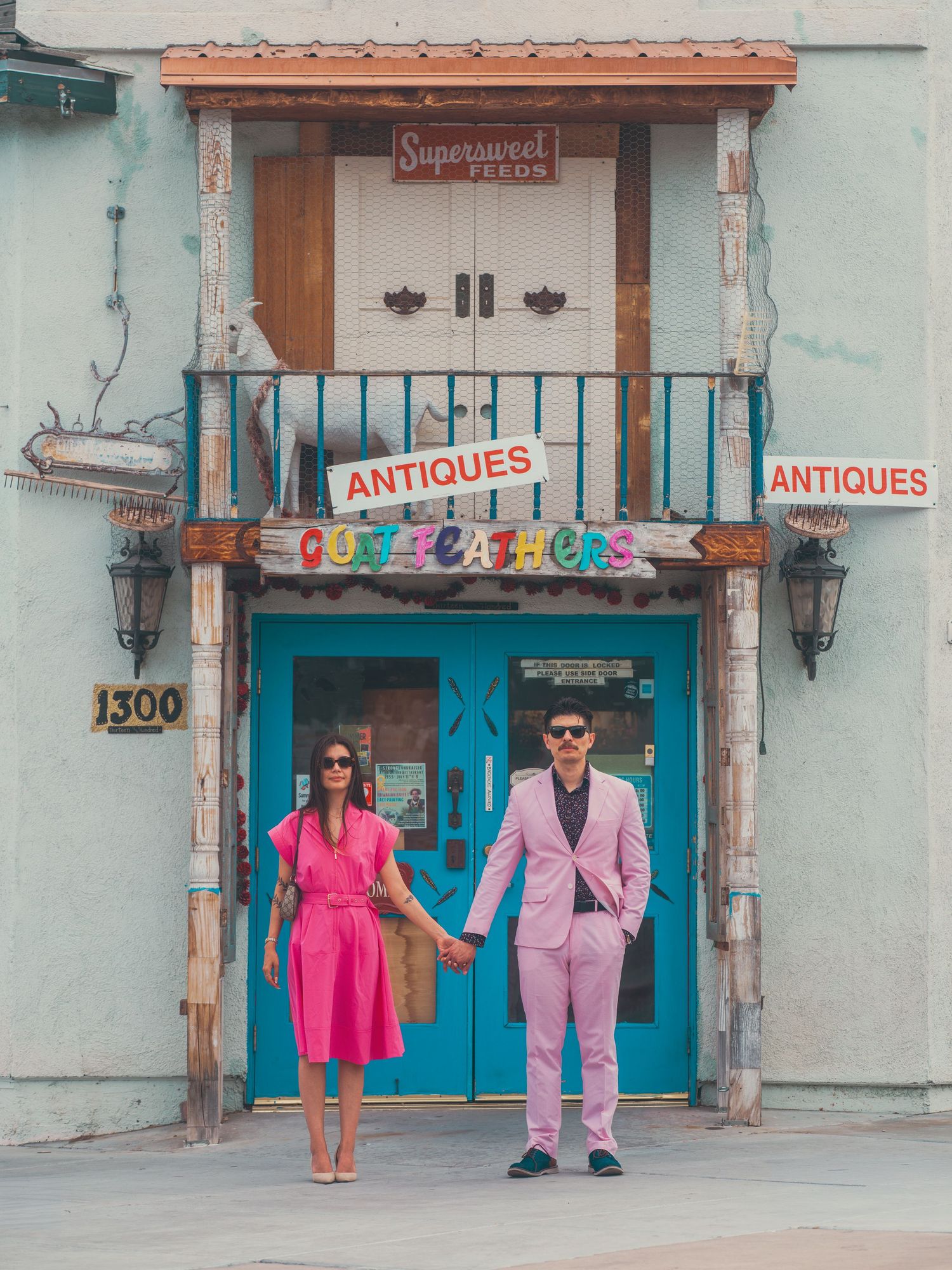 Couple in matching pink outfits holding hands outside a colorful antiques shop with a turquoise door.